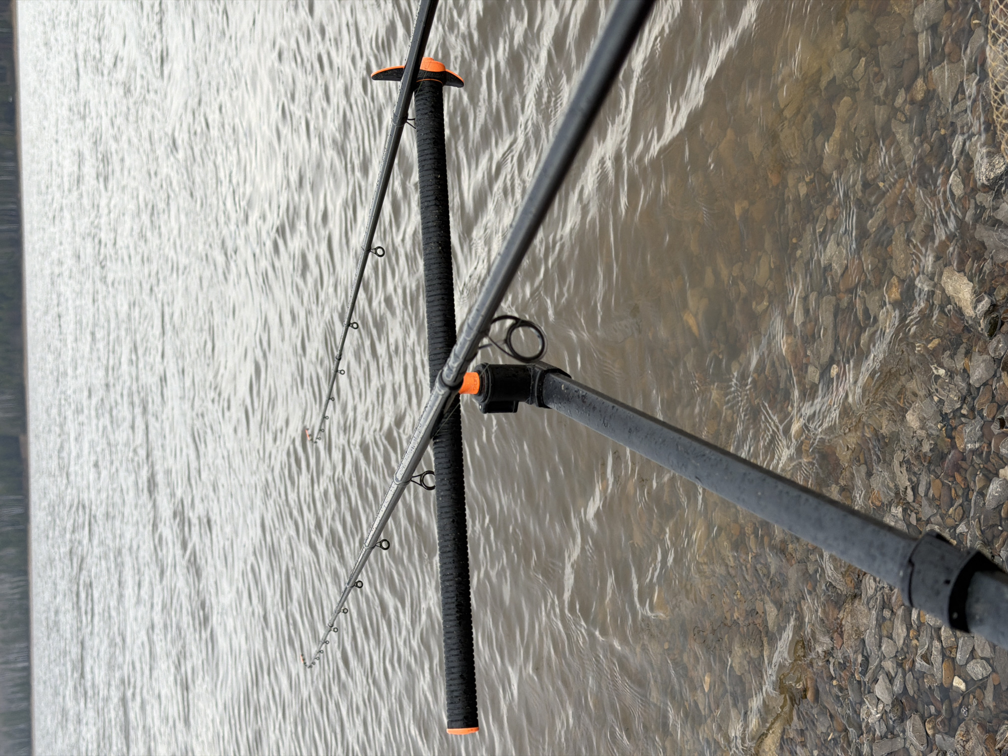 Multiple black fishing rods with bright orange accents are positioned diagonally across the frame, indicating an active fishing setup. One prominent rod extends from the lower right towards the upper left, while another is visible largely on the left side. These rods are supported by a robust, black, ribbed structure, likely a rod pod or bank stick system, positioned vertically towards the center-right.
The left and central parts of the image are dominated by a body of water, possibly a river or lake, displaying numerous fine, horizontal ripples across its surface, suggesting movement or a gentle breeze. The right side of the frame features a shallower area where the water is clear, allowing a detailed view of the rocky and pebbled bed beneath. The overall lighting is soft and diffused, typical of daytime conditions, potentially under an overcast sky, without harsh shadows. The scene captures the equipment and environment of an angler, focusing on the fishing gear set by the water's edge. No specific time of day or emotional expressions are discernible, and no text is visible within the image. This image was taken in Chaikovsky, Russia.