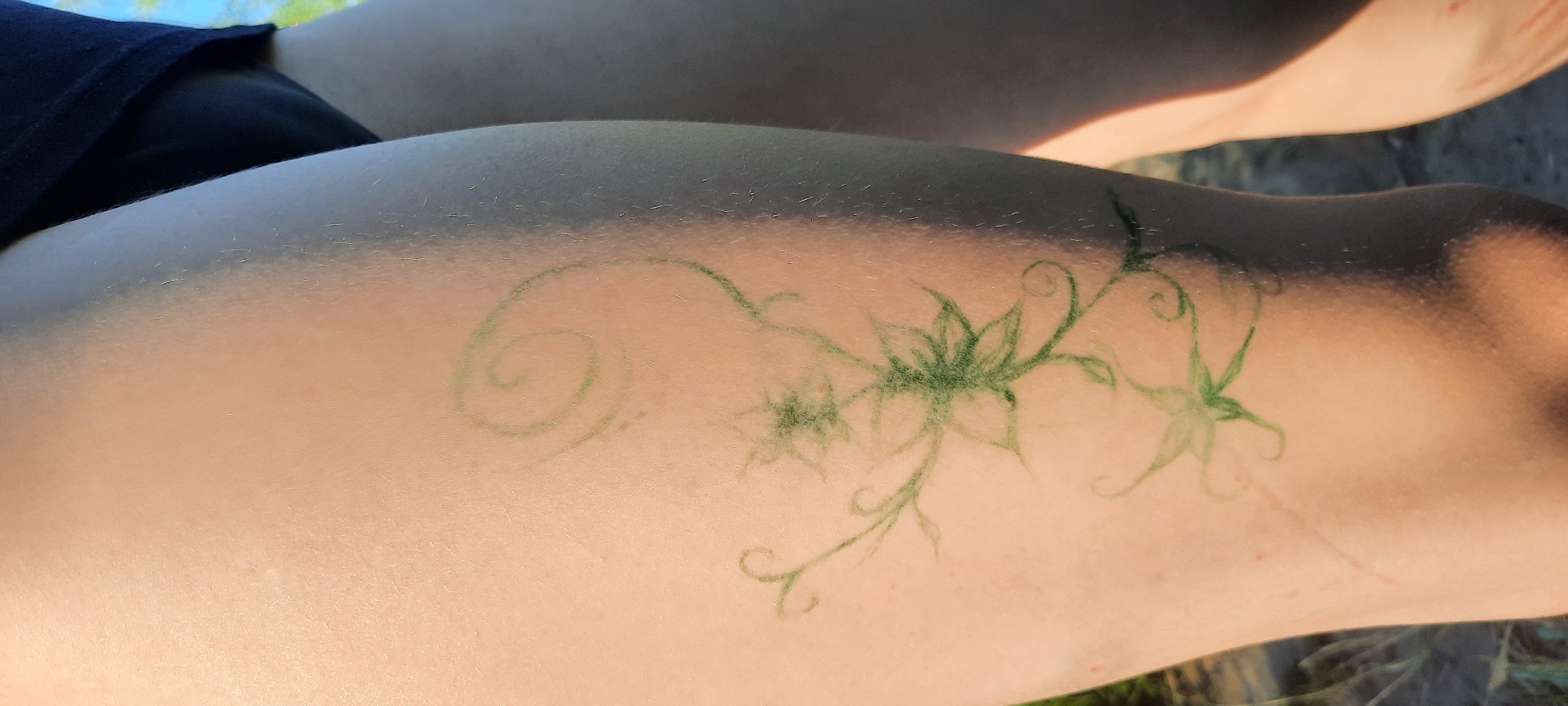 A close-up shot focuses on a person's thigh, where a delicate, green, henna-like tattoo of flowers and vines is visible. The tattoo is positioned on the upper part of the thigh, extending horizontally across the frame. The skin appears tanned with some areas lighter due to shading. A dark blue garment, possibly shorts, is visible at the top left of the image. The background is blurred, suggesting an outdoor setting with greenery and possibly sunlight filtering through trees, indicating a daytime scene. The overall impression is one of a casual outdoor activity. The tattoo's artistic design and the natural lighting contribute to a serene and summery ambiance. There is no visible text in the image.