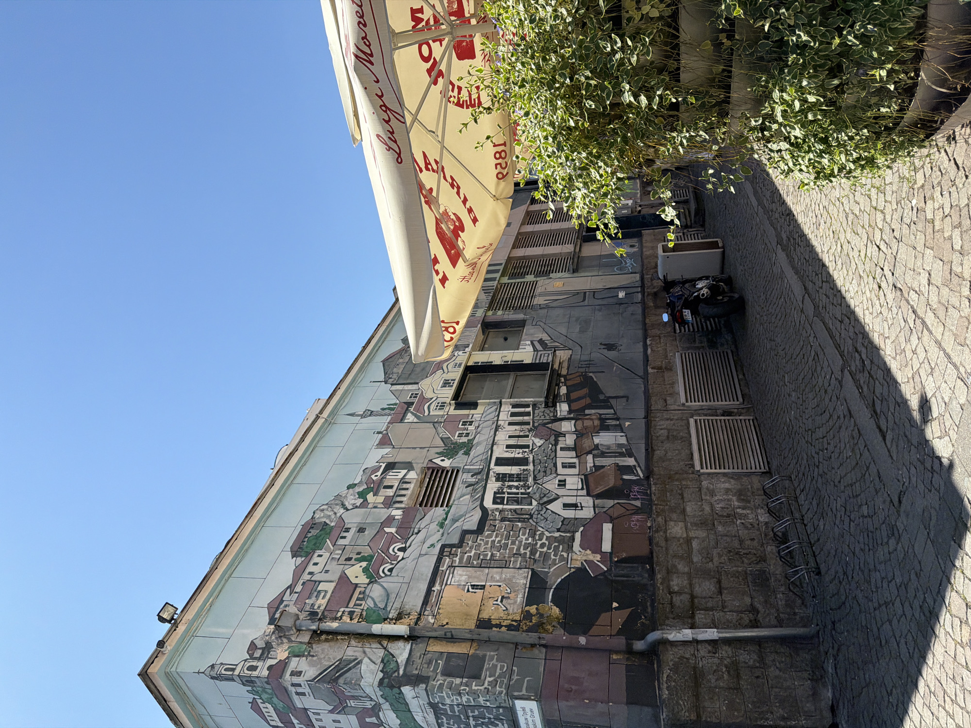 The image captures a street scene in Plovdiv, Bulgaria, focusing on a building with a large, detailed mural depicting a stylized cityscape. The mural spans the side of the building and extends from near ground level to the roof. Above the mural, clear blue sky is visible.
A large patio umbrella with the "Birra Luigi Moretti" logo and advertisement in red dominates the upper right corner of the image, partially obscuring the building behind it. Green foliage hangs down from a planter, further obscuring the building and the street.
On the street, a parked motorcycle is visible, along with a row of metal bicycle racks attached to the building. The street is paved with cobblestones, and a shadow stretches across it, suggesting strong sunlight from an unseen direction. The overall atmosphere is bright and sunny, with the focus on the mural and the elements framing it.