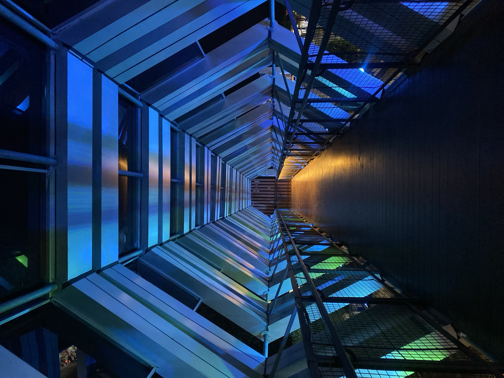 The image shows a pedestrian bridge, lit with blue and green lights. The bridge has a walkway made of dark wood and metal railings. The ceiling of the bridge is made of metal beams and panels, which are also illuminated with blue and green lights. The lights create a tunnel effect, drawing the viewer's eye towards the end of the bridge, where there is a wooden gate. The scene is set at night, and the overall mood is futuristic and somewhat mysterious. The location of this pedestrian bridge is the city of Banjarbaru, Indonesia, specifically in the area of the Banjarbaru Great Mosque.