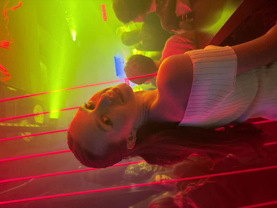 A young woman with dark hair and light skin poses in a nightclub setting. She wears a white, ribbed top and dangling earrings. The background features blurred figures and bright, colorful lighting including pink horizontal lines and yellow light. Her face is slightly tilted, with a neutral expression. The overall atmosphere is vibrant and energetic.