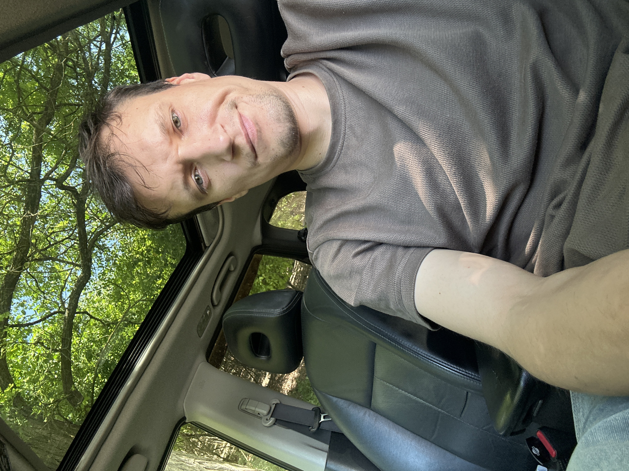 In Makiv, Ukraine, a man takes a selfie inside a car with a sunroof open, revealing a canopy of green trees above. He is seated in the driver's seat, wearing a brown t-shirt. The car interior features black leather seats and a seatbelt. The scene appears to be a still moment, with the man looking towards the camera with a slight smile.