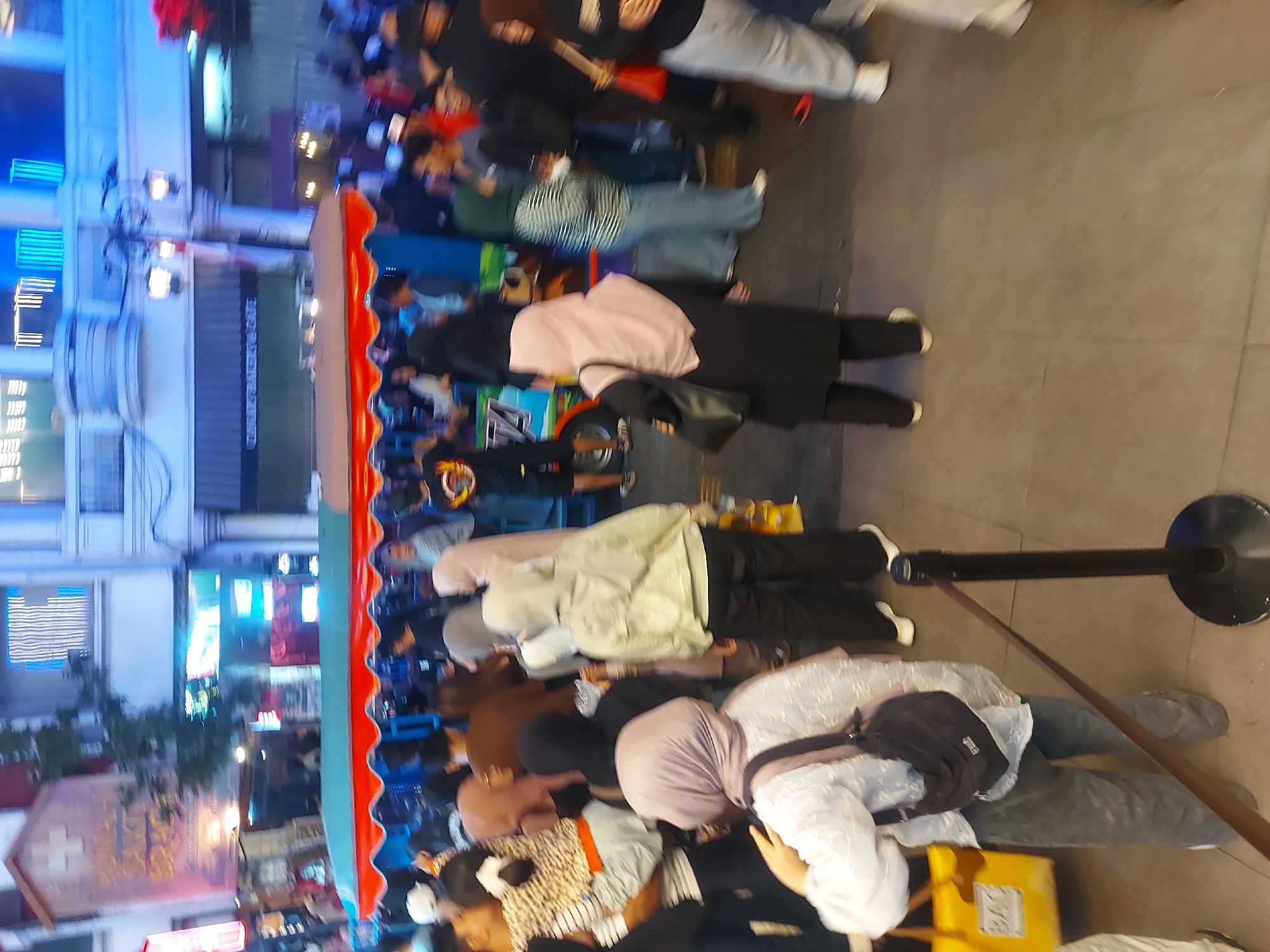 The image shows a bustling street scene in Bandung City, Indonesia, likely in the evening given the lighting. A brightly colored tram-like vehicle, possibly a mobile food or souvenir stand, is central to the composition. Numerous people, many wearing headscarves, are gathered around the vehicle and walking along the paved street. The architecture in the background suggests a commercial area, with storefronts and illuminated signs, some featuring what appear to be blue neon lights. The overall atmosphere is dynamic and crowded, indicative of a popular public space.