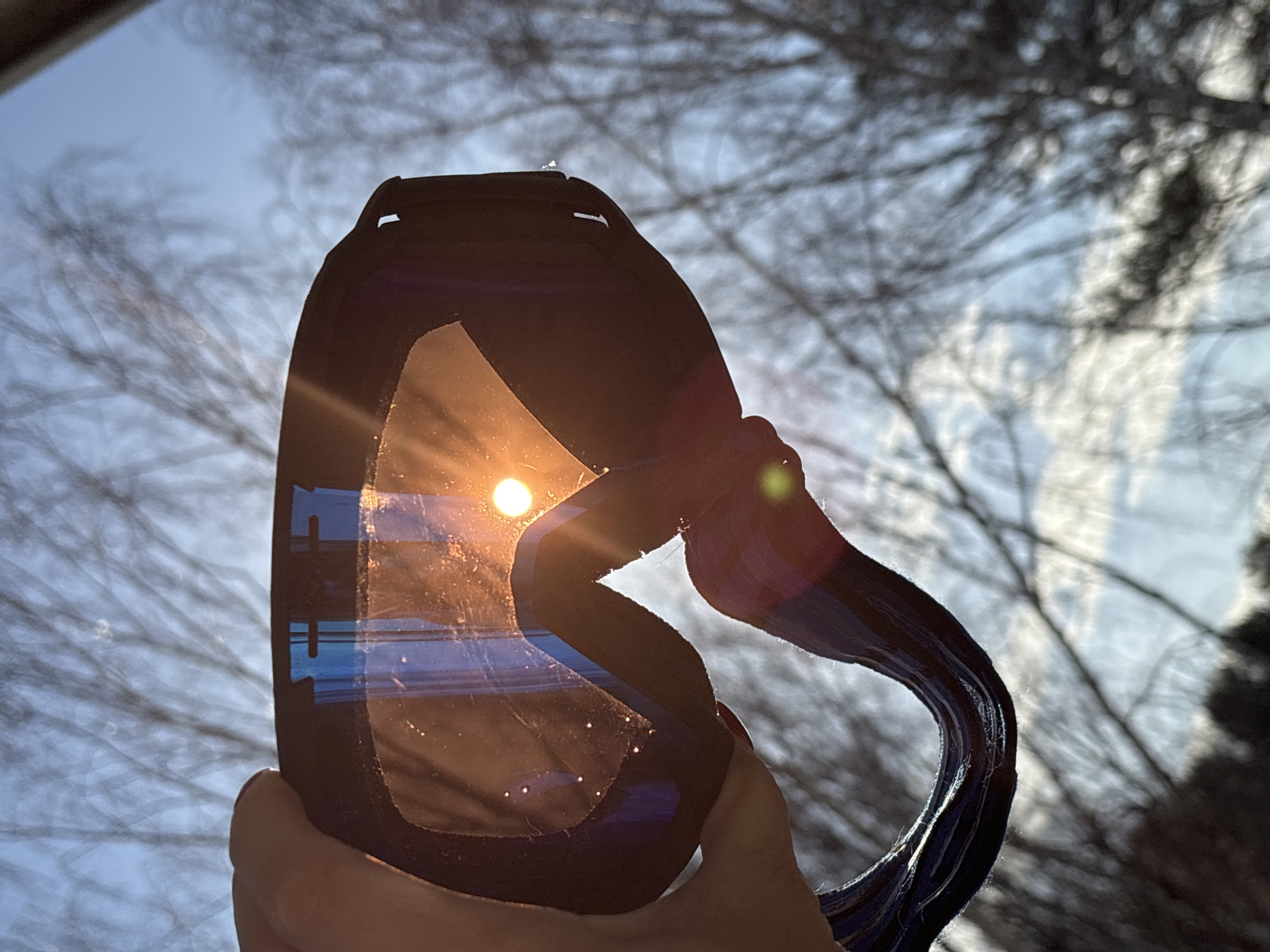 The scene is set outdoors in Chaikovsky, Russia. A human hand, with a visible red-painted fingernail on the thumb, grips the upper-left edge of a pair of dark-framed ski or snowboard goggles. The goggles' lens, which appears to have a slightly tinted or reflective surface, is positioned directly in front of the sun, which manifests as a brilliant, circular orange-yellow orb located slightly right of the lens's vertical center. The lens exhibits multiple small specular highlights and scattered dust or scratch marks. Reflections of blue light are visible on the upper and lower portions of the lens. The dark blue or black goggle strap extends downwards from the right side of the frame, partially visible and out of focus. The background consists of a heavily blurred array of bare tree branches silhouetted against a light blue sky, indicating an environment likely experiencing autumn, winter, or early spring. The sun's angle suggests either early morning or late afternoon.
