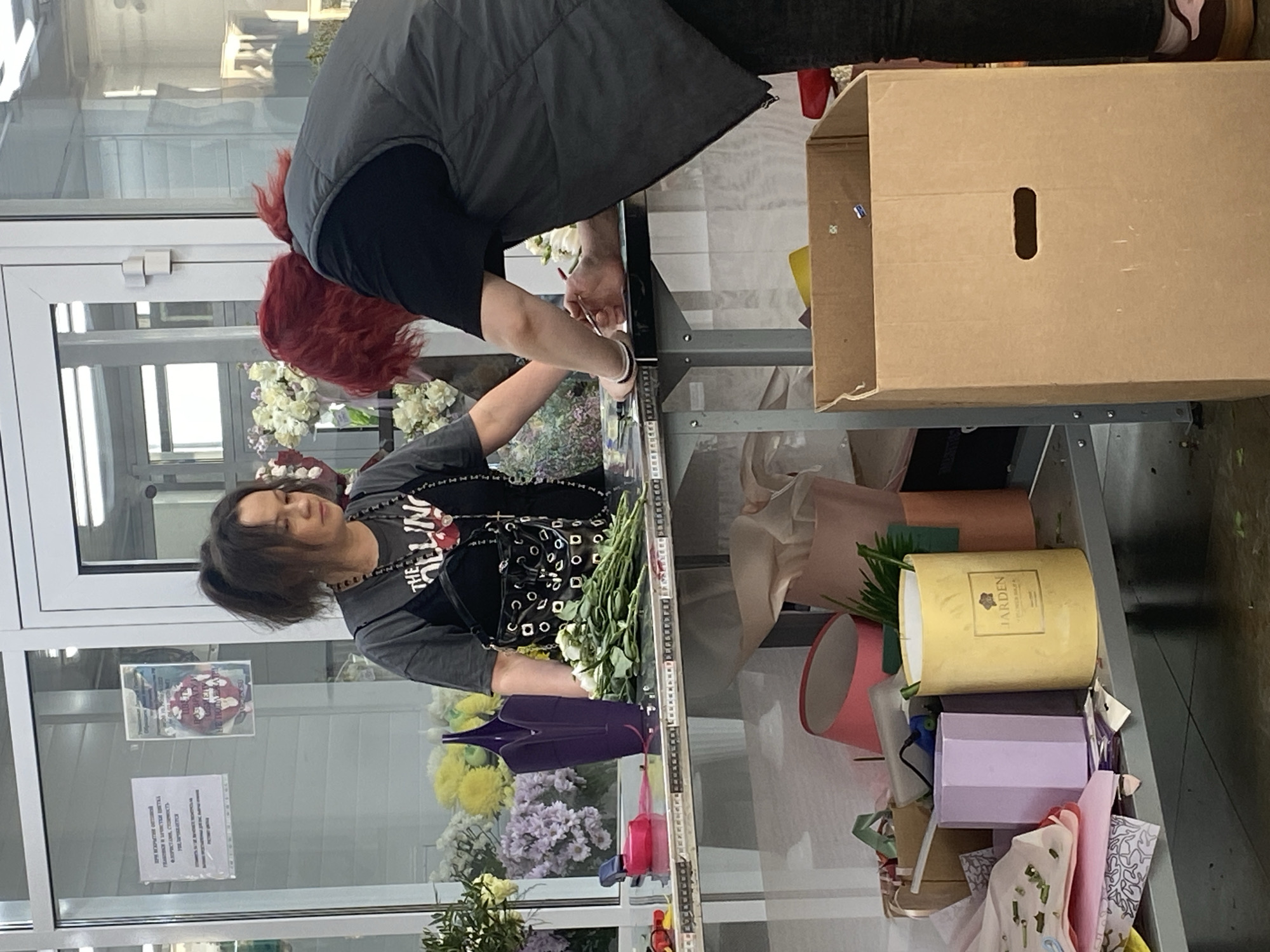 Two individuals are arranging flowers on a glass-topped counter in what appears to be a flower shop. One person has red hair and is wearing a grey vest, while the other person has dark hair and is wearing a grey t-shirt. The counter is laden with various flowers and floral arrangements, alongside gift boxes and supplies. A large cardboard box sits to the right of the counter. Through the shop window, a poster and informational signs are visible. The location is identified as Yekaterinburg, Russia.