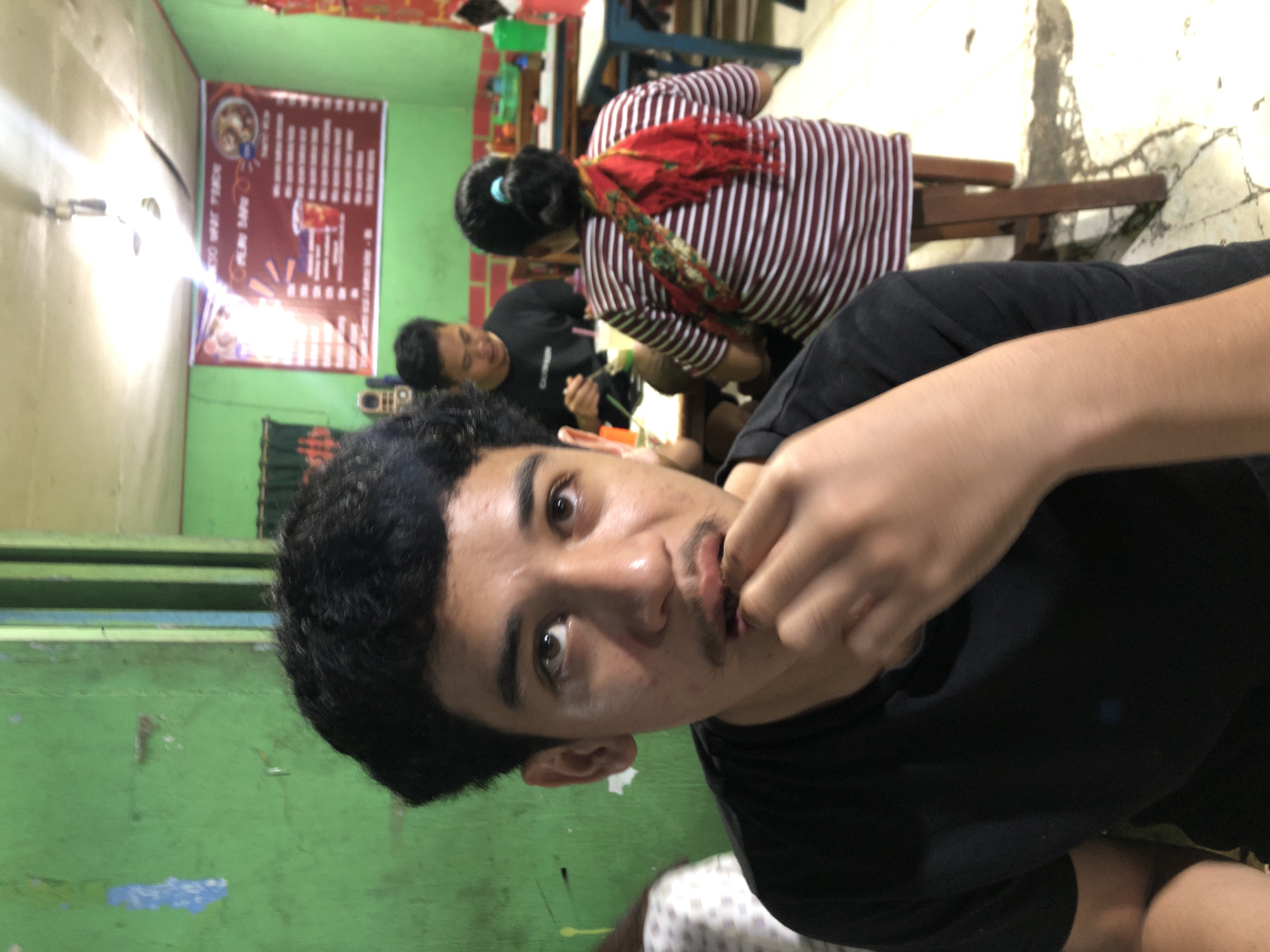 The image depicts a group of people in what appears to be a casual eatery in Sei Mencirim, Indonesia. In the foreground, a young man with dark, curly hair and a mustache is eating, with his hand near his mouth. He is wearing a black t-shirt. Behind him, a man wearing a black t-shirt is seated, looking down at his food. To the right, a woman with her hair in a bun and wearing a red and white striped shirt and a colorful scarf around her neck is also seated. The environment features green walls, a banner with text and images on the wall, a ceiling fan, and tables and chairs. The lighting is dim, with a bright light source above, possibly a lamp. The floor appears to be tiled.