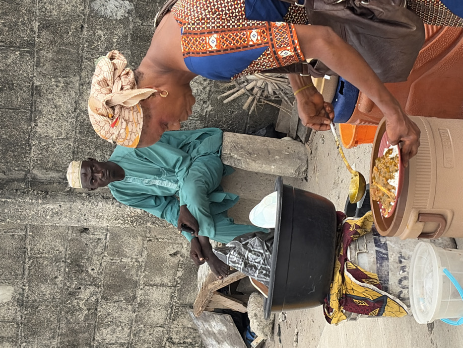 An outdoor scene in Jakande, Nigeria, features two individuals and an informal food vending operation. In the foreground, a woman with dark skin and a patterned beige headwrap is actively serving food. She wears a blue top with patterned orange and brown sleeves and a brown shoulder bag. Her right hand holds a golden ladle, scooping from a container, while her left hand supports a red plate containing white rice topped with a brown stew with meat pieces. The plate rests on a beige "Pot Trendy" insulated food container, adjacent to a blue container. To her left, a large black plastic basin filled with miscellaneous items, including a black plastic bag, sits atop two stacked white/grey buckets draped with a yellow and red patterned fabric. A clear plastic bucket is visible on the ground.
In the background, a man with dark skin and a teal-colored traditional garment (tunic and trousers) is seated barefoot on a concrete block. He wears a light-colored embroidered cap and has a visible mark on his forehead. His hands are clasped in his lap, possibly holding a small dark object. He looks directly forward with a serious expression. Behind him is a textured, weathered grey concrete block wall. The ground is uneven, composed of dirt and debris, with a wooden stool and some scattered sticks visible near the man.