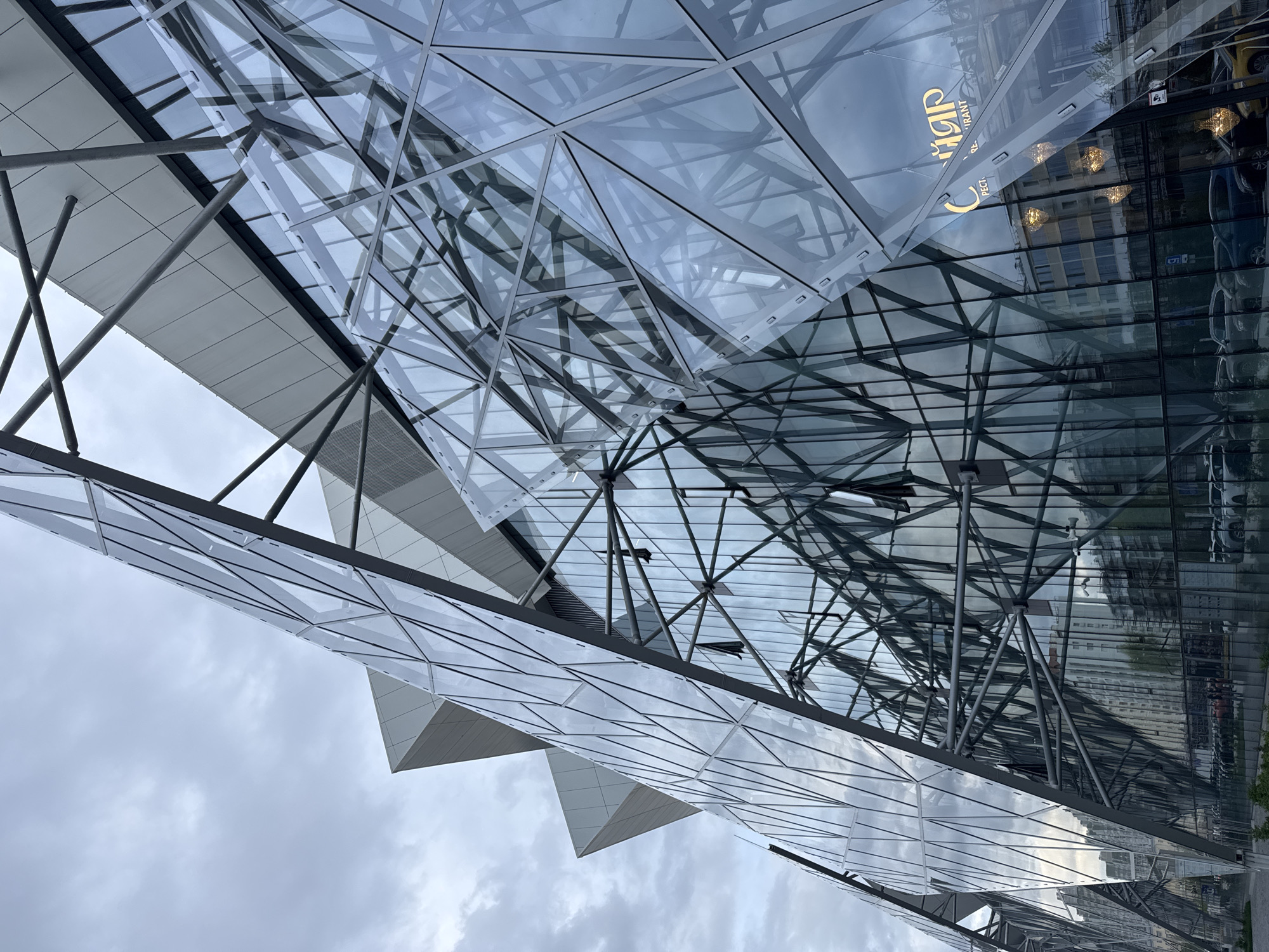 The image captures a section of the "Kazan" restaurant in Kazan, Russia, specifically focusing on the complex architectural design of its exterior. The restaurant exhibits a modern design, characterized by interconnected triangular glass panels that create a geometric, almost crystalline appearance. The glass reflects the sky, clouds, and surrounding buildings. Steel support beams reinforce the structure. The foreground shows the sky. The image provides a close-up perspective of the building's facade, emphasizing its unique architectural features.