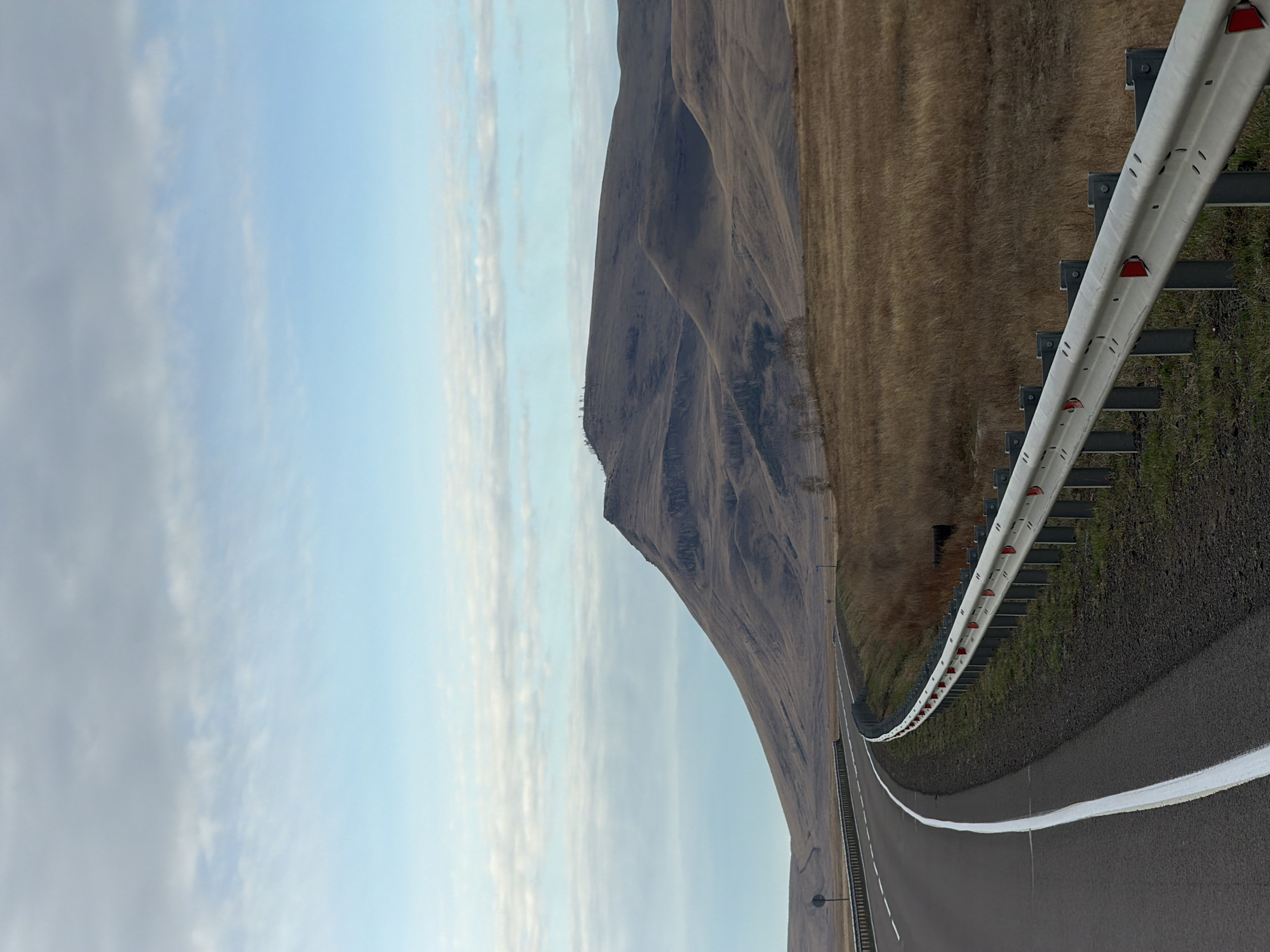 A landscape scene in or near Сарагаш, Russia. The image shows a paved road curving along the base of a tall, barren mountain. To the road's right, a metal guardrail is visible. Beyond the guardrail is a field of brown grass. To the left, the sky is light blue with thin, wispy clouds. The road's surface is dark asphalt with white lane markings.