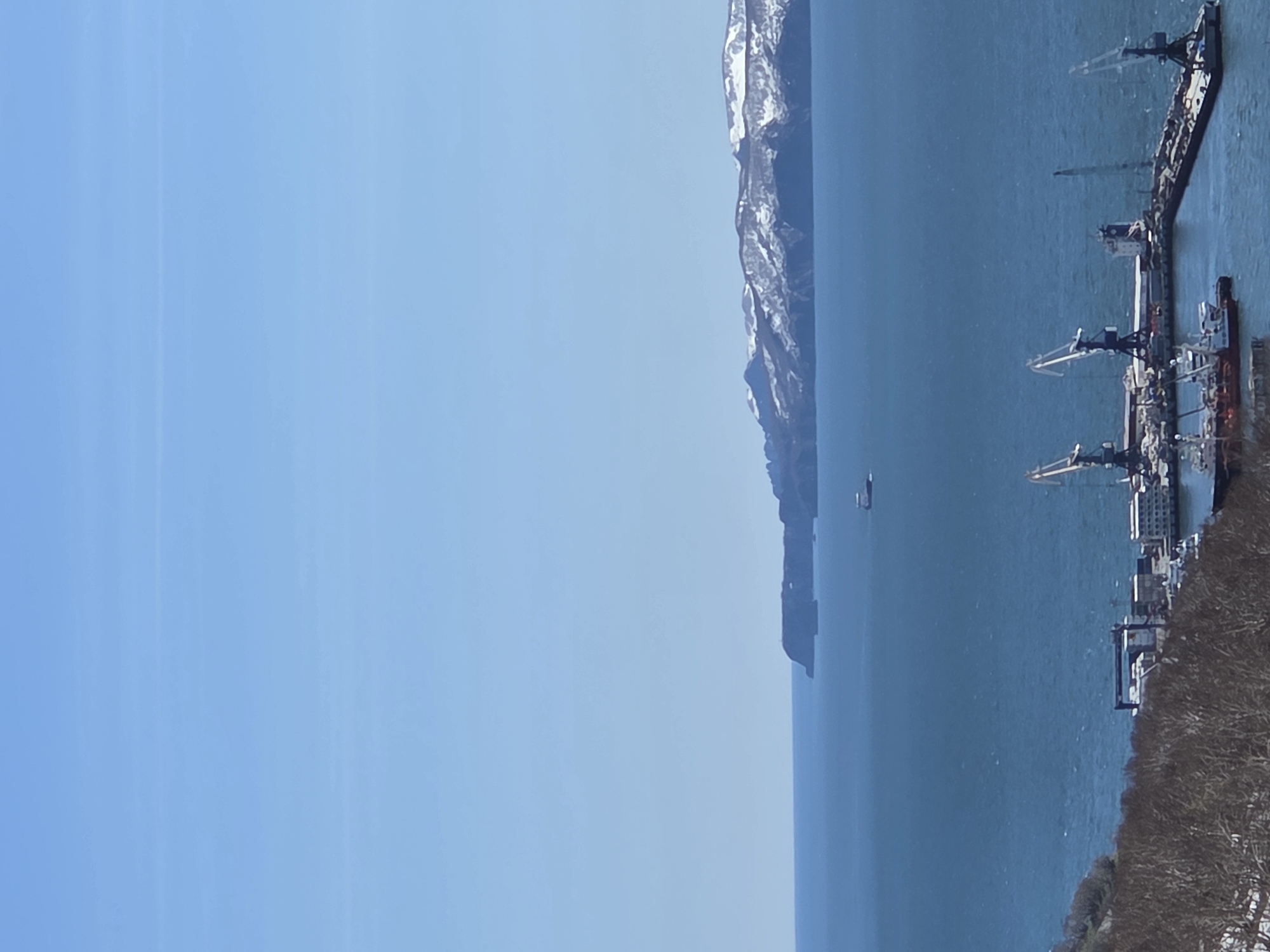 The image depicts a coastal scene. A strip of land or peninsula with snow-capped mountains extends into the water. In the foreground, there is a pier or dock area with several ships and cranes. The sky is clear blue. The exact location and city are not discernible from the visual information provided.
