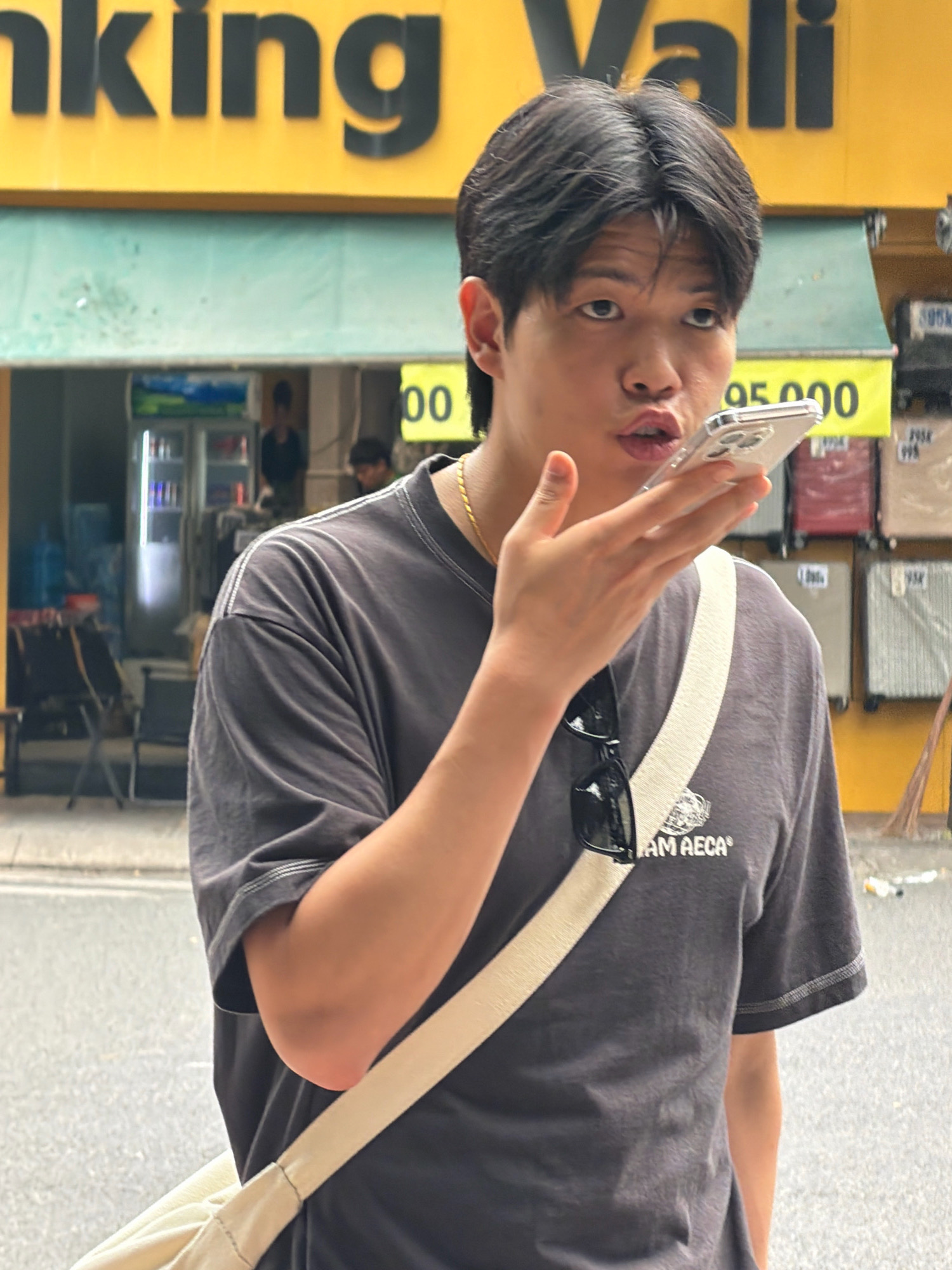 The image shows a young East Asian man holding a smartphone to his ear, appearing to be on a call, in front of a yellow building with the word "anking" and "ali" visible on it, suggesting it is a bank. He is wearing a dark grey t-shirt with a white strap of a bag across his chest and sunglasses hanging from the neckline. Behind him, a turquoise awning covers a storefront with a refrigerator visible inside. In the background, there are stacks of luggage and a yellow sign with the numbers "95.000". The scene appears to be on a street in Nha Trang, Vietnam, indicated by the context of similar images. The man's expression is one of active listening or speaking.