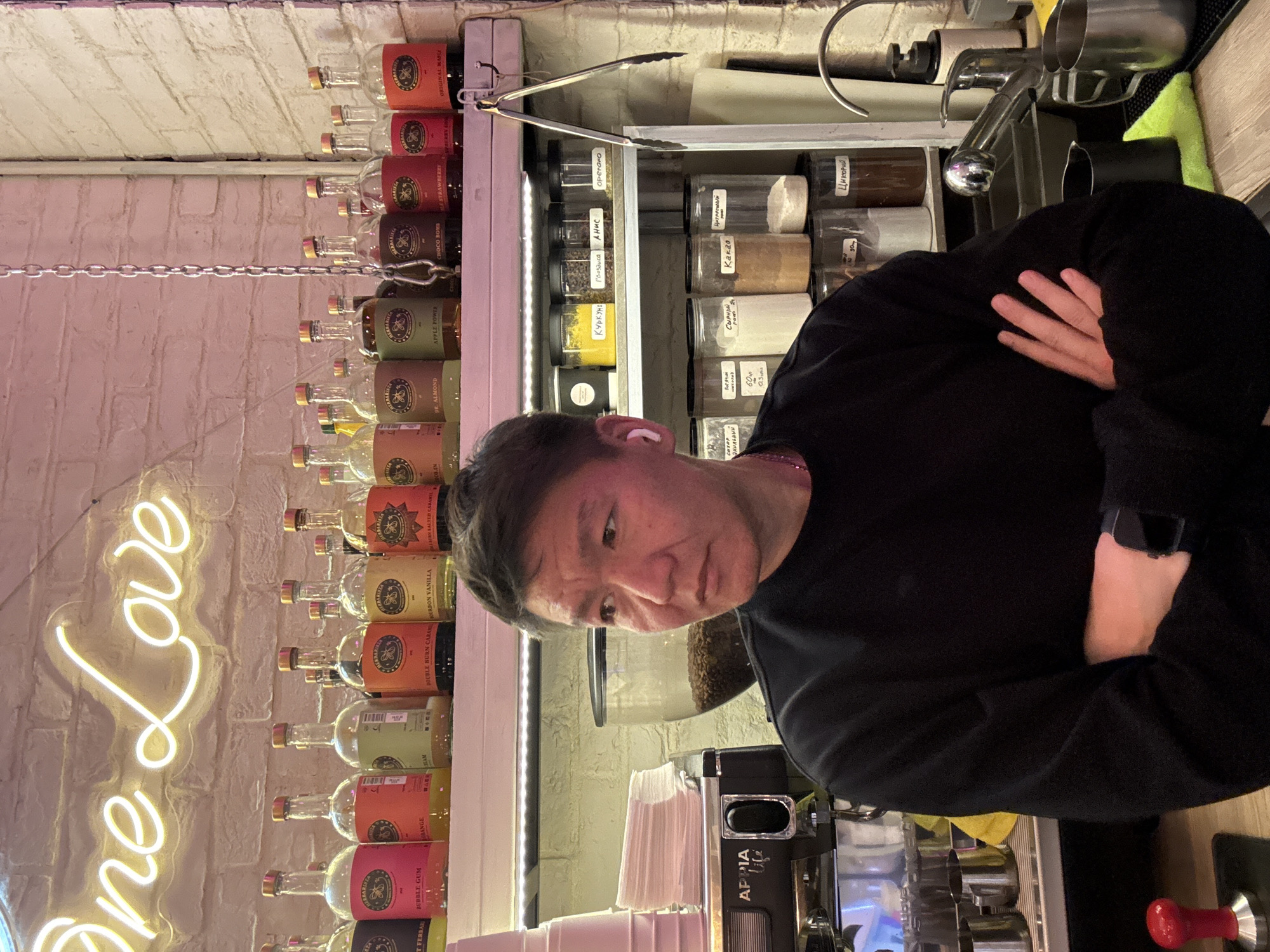 A medium shot captures a man with dark hair, wearing a black long-sleeved shirt, standing with his arms crossed. He is positioned in front of a shelf stocked with various glass bottles, all featuring red, yellow, and green labels with the brand "HERBARISTA" visible. Above the bottles, a neon sign spells out "One Love" in cursive white lettering against a brick wall. To the man's left, on a lower shelf, are stacks of white paper cups. Behind him, more shelves are filled with clear jars containing various ingredients, some labeled in Russian script. A coffee machine is visible on the counter to the left, and a faucet and sink are to the right. The scene appears to be a cafe or bar environment in Moscow, Russia, indicated by the Russian labels on the jars.