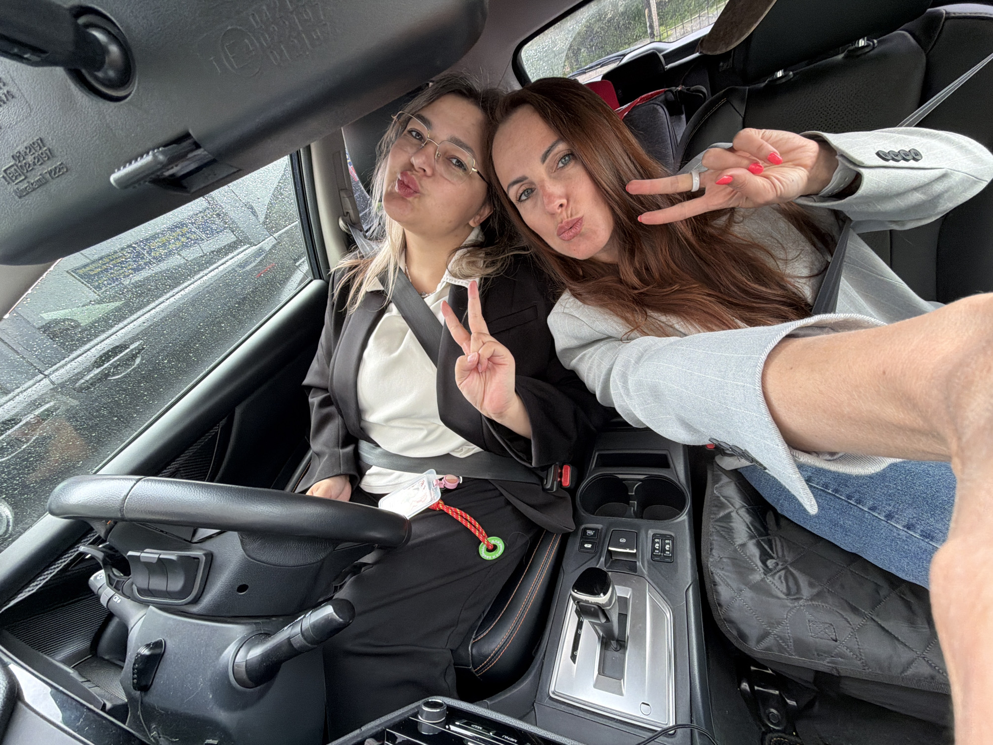 This is a selfie taken inside a car, featuring two women posing playfully. The woman on the left, with light brown hair and glasses, is wearing a black blazer over a white top and a seatbelt. She is making a peace sign with her left hand and puckering her lips. Attached near her seatbelt buckle is a small white plastic container and a green circular tag with a red and yellow braided rope. The green tag has text in Cyrillic: "НАСТОЯЩЕЕ", "БИРОБИДЖАН", and "С ЛЮБОВЬЮ".
The woman on the right, with long brown hair, is wearing a light grey pinstriped blazer and blue jeans. She is also puckering her lips and making a peace sign with her right hand, which has red nail polish and a white ring on her middle finger. Both women appear to be in good spirits, expressing a friendly and cheerful mood.
The interior of the car is visible, including the black dashboard, steering wheel, gear shifter, and cupholders. A black quilted seat cover is on the passenger side. Water droplets are visible on the car windows, suggesting it is raining or has recently rained. Reflections of the exterior environment, possibly another vehicle or structures, can be seen in the wet windows. The rearview mirror overhead has visible text and numbers: "E" (within a circle), "045197", "102197", "01.2197", and "Miracom T225". The scene appears to be set during the daytime, likely under overcast conditions.