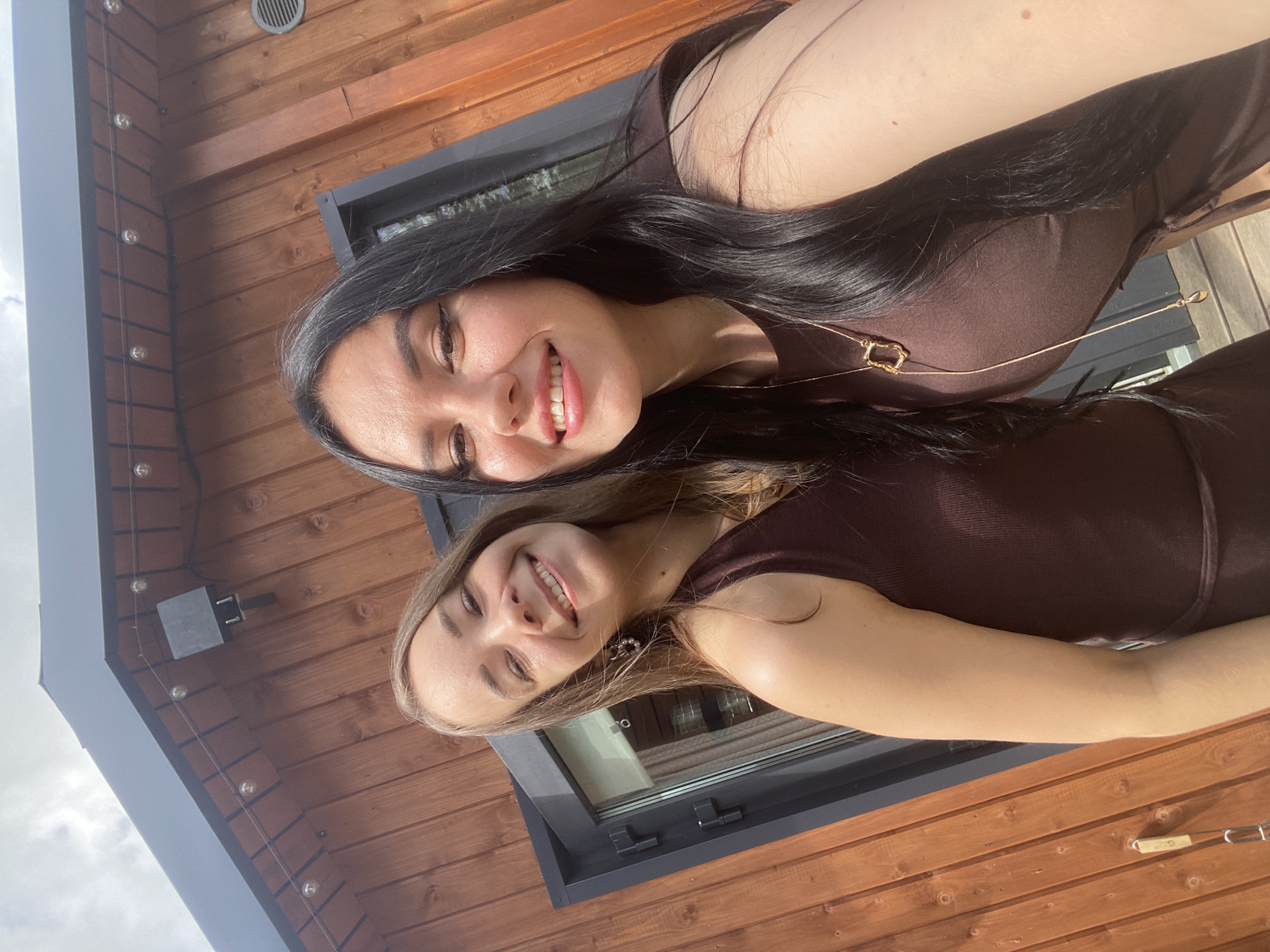 Two young women, one with long dark hair and the other with long blonde hair, pose for a selfie outdoors. They are both wearing brown sleeveless dresses. The woman with dark hair has a gold necklace with a pendant. Behind them is a wooden building with a dark-colored roof and a black framed window. String lights are visible under the roof eaves. The scene appears to be in a residential or recreational area, possibly a dacha or a private home. The sky is overcast with some patches of blue. There are no specific identifiable landmarks that place this image in Chaikovsky, Russia, beyond the information provided.