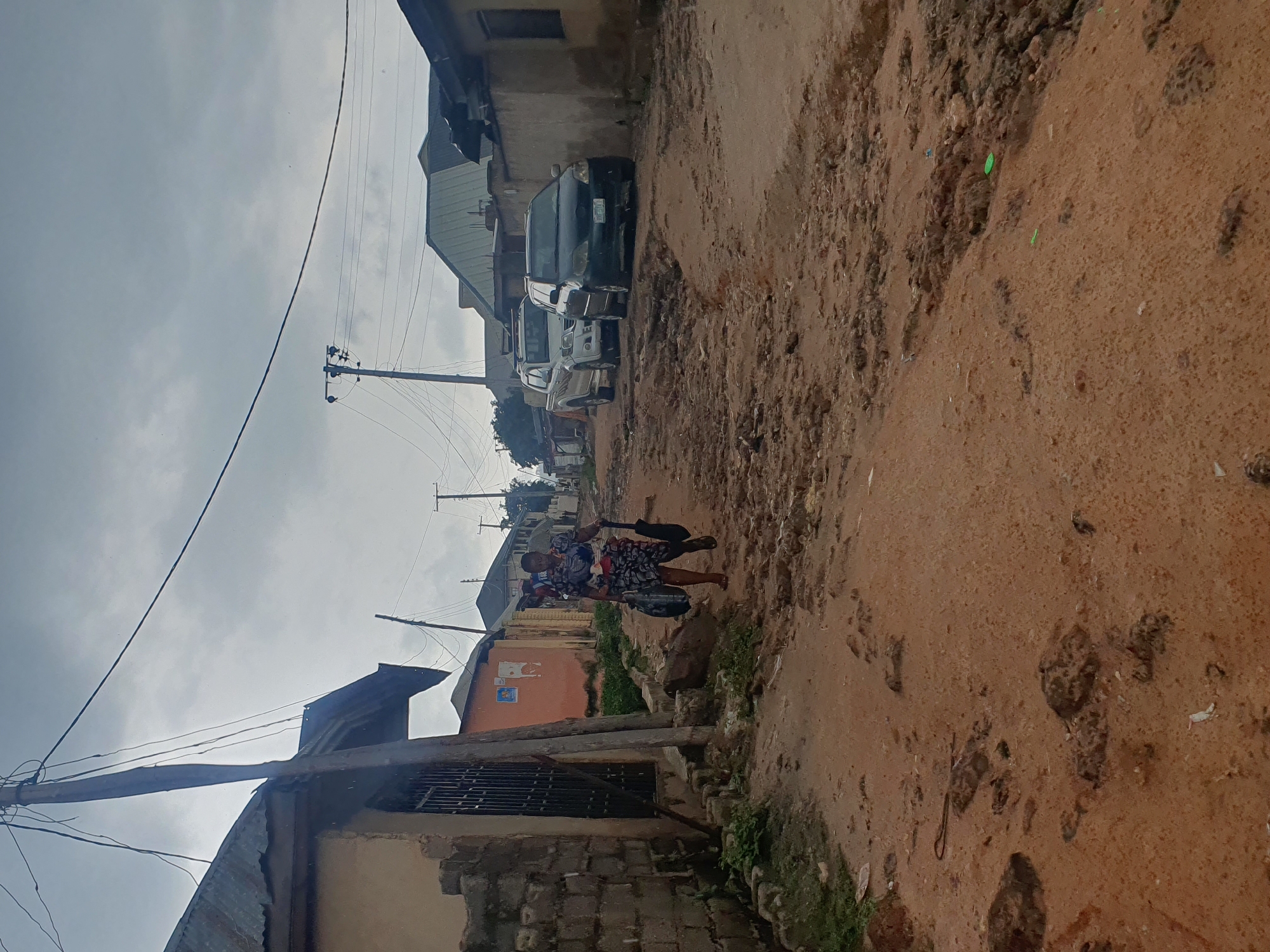 A woman walks down a muddy, unpaved road in Lokoja, Nigeria, carrying two large black bags and a black umbrella. The road is in a state of disrepair, with large puddles and uneven terrain. On either side of the road are houses with corrugated metal roofs and bare brick walls. A few vehicles, including a white pickup truck and a dark sedan, are parked on the road. Overhead, power lines crisscross the cloudy sky. The overall impression is one of a rural or semi-urban environment in need of infrastructure development.