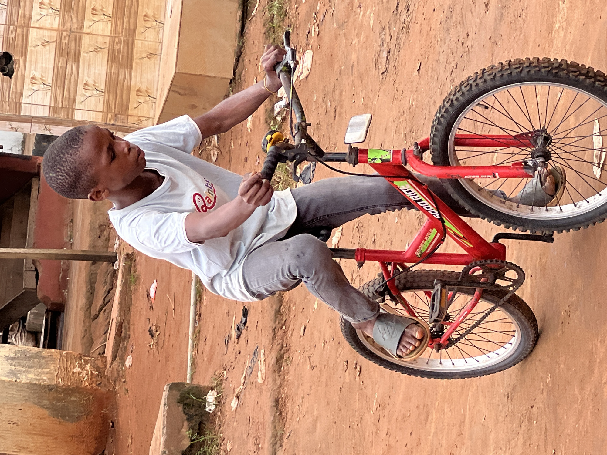 A young boy rides a red bicycle on a dirt road in Nkpor, Nigeria. The boy is wearing a light blue t-shirt and grey pants. The bicycle has black tires and a red frame. The background consists of a dirt road, some buildings, and vegetation. The image is taken from a medium distance, capturing the boy and his bicycle in motion. The lighting suggests it is daytime, with natural light illuminating the scene. The overall impression is one of everyday life in a Nigerian urban setting.