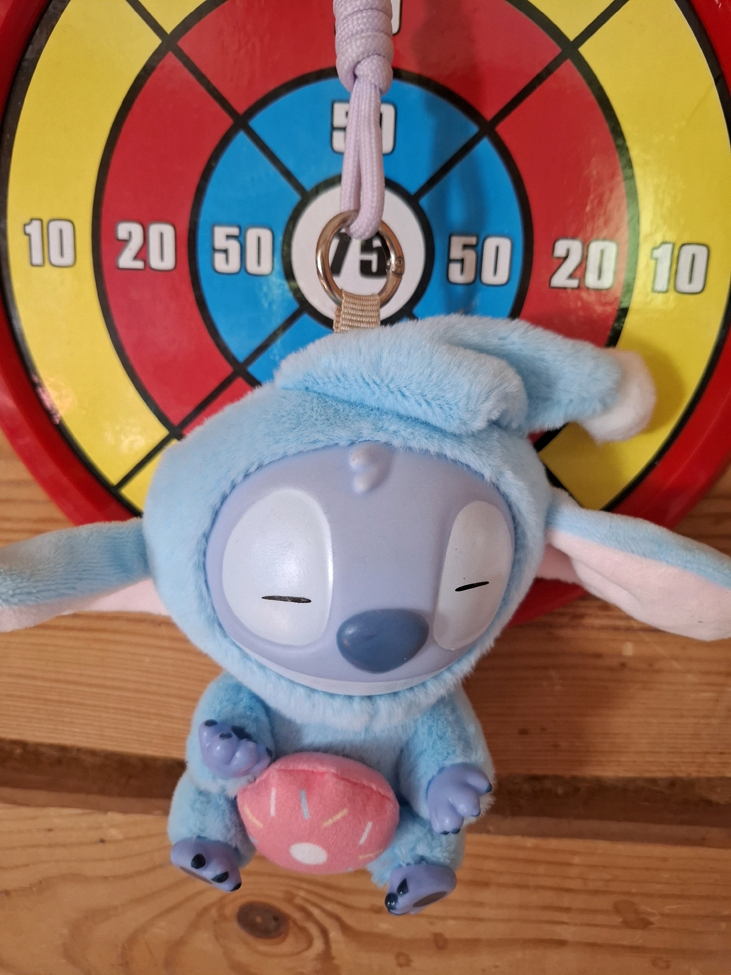 The image shows a blue plush toy resembling the character Stitch, wearing a blue sleeping cap. The toy is sitting in front of a circular dartboard with concentric rings of yellow, red, and blue. Numbers 10, 20, and 50 are visible on the dartboard. The toy is holding a pink donut-shaped object with white sprinkles and a white center. A light purple cord with a metal ring is attached to the top of the toy. The background appears to be wooden planks. The image does not contain information about the city of Tyumen, Russia, or any specific location.