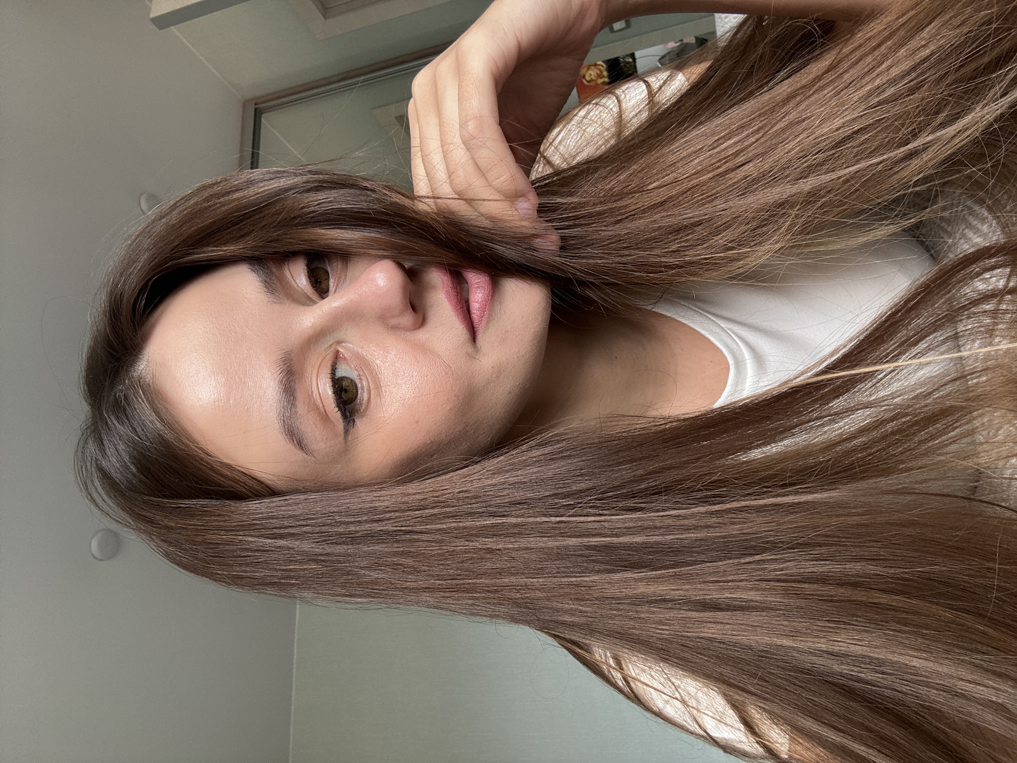 A woman with long, brown hair is posing indoors, likely in a residential setting. She has fair skin, brown eyes, and is wearing lip gloss. She is wearing a white t-shirt. The background consists of a light gray wall and a window or a mirrored closet door. The woman's hand is near her face, partially obscuring it with her hair. The overall lighting appears soft and natural. It is impossible to determine the location in the city of Krasnoyarsk, Russia from the image.