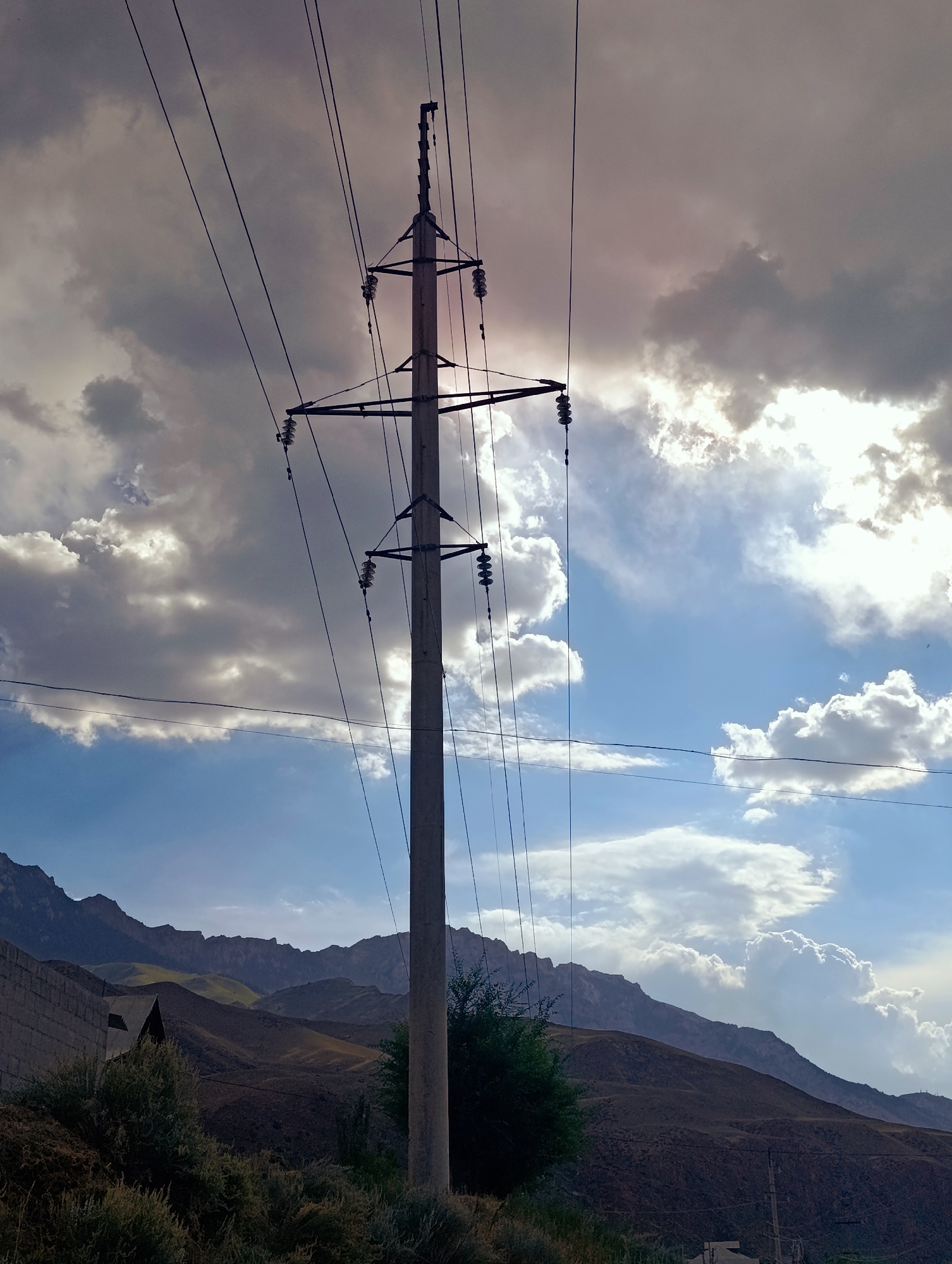 An outdoor, eye-level shot captures a concrete utility pole with a network of electrical wires against a dramatic sky filled with dramatic clouds and patches of blue. The sun is partially obscured by the clouds, creating a backlit effect. In the background, rugged, brown-toned mountains rise, with hints of green vegetation on their slopes. In the foreground, to the left, a section of a grey brick wall and dense, dark green bushes are visible. The overall scene suggests a rural or mountainous environment near Naryn City, Kyrgyzstan.