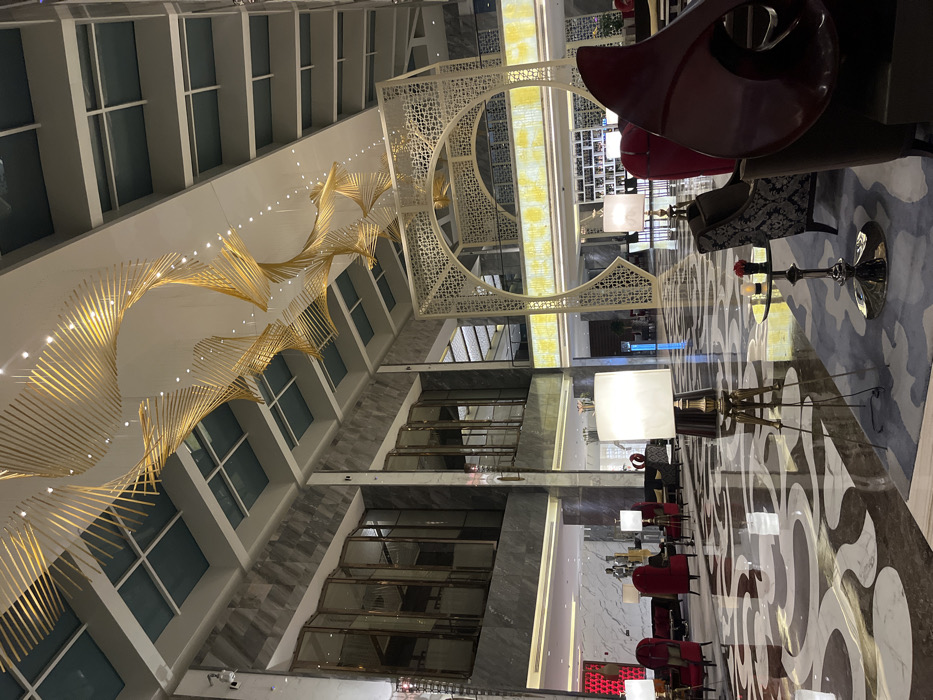 This image depicts a vast, high-ceilinged interior space, identified as being within Yanta District, China, consistent with a modern hotel lobby or grand public area. The polished marble floor displays a distinct, sprawling pattern of dark brown and off-white organic shapes, with a blue patterned rug visible in the immediate foreground on the right.
The ceiling features multiple structural levels, recessed lighting fixtures, and several rectangular skylights with a greenish hue. A prominent, large golden chandelier dominates the upper central space, constructed from numerous thin, metallic, spiraling elements that form an elongated, wave-like sculptural array.
Walls are clad in polished grey marble or stone panels. On the left side of the frame, an upper level showcases a series of tall, segmented architectural panels or windows with dark frames, beneath which an illuminated horizontal strip is present. To the right, a large, ornate, light-colored archway or screen, characterized by intricate geometric lattice work, serves as a decorative focal point or partition, extending upwards as part of a multi-story structure. A brightly illuminated horizontal band, emitting a yellow-toned light, is situated below the upper sections of this archway.
On the ground level, various seating arrangements are distributed. In the foreground right, a dark, patterned armchair is partially visible next to a small, polished silver pedestal table holding two dark, cylindrical objects. Two tall floor lamps with square, illuminated white shades and dark bases are positioned in the mid-ground. Multiple plush, high-backed red velvet armchairs with gold-colored accents are arranged on the left side, some accompanied by small side tables. Additional dark upholstered seating is visible further in the background. No individuals are present within the scene.