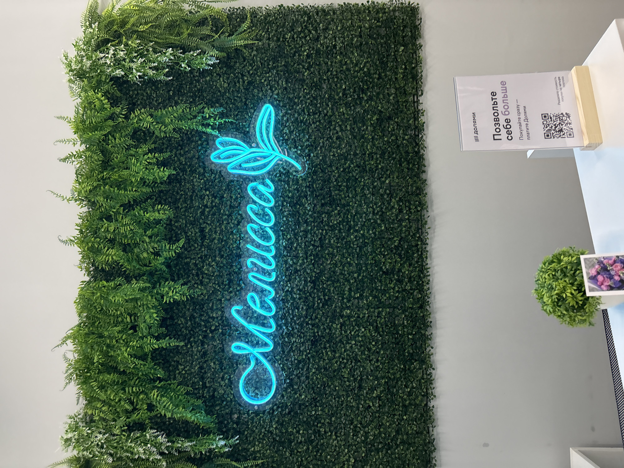 An indoor scene within the "Melissa" beauty salon located at Karl Marx Street, 32, Chaikovsky, Perm Krai, Russia. The focal point is a large artificial green wall, densely covered with small, dark green foliage, from which larger artificial ferns and white-flowered greenery cascade from the top. Centered on this green wall is a bright blue neon sign in cursive Cyrillic script, spelling "Мелисса" (Melissa), followed by a stylized plant or leaf motif.
Below the green wall, a clean white counter or reception desk is visible. On the left side of the counter, a small potted artificial plant with vibrant green, dense foliage rests in a white pot, next to a small white frame displaying an image of pink and purple flowers. On the right side of the counter, a clear acrylic sign holder with a light wooden base presents a vertical white sign. The sign features the "III долями" (III Doliami) logo at the top and the prominent Russian text: "Позвольте себе больше" (Allow yourself more), with smaller text below reading: "Покупайте сразу – платите Долями" (Buy now – pay with Doliami). A QR code is present on the sign, underneath which smaller text states: "Разделите стоимость покупки на четыре платежа" (Divide the purchase cost into four payments), indicating a "buy now, pay later" service. The environment is well-lit and appears clean and contemporary. No individuals or active interactions are depicted.