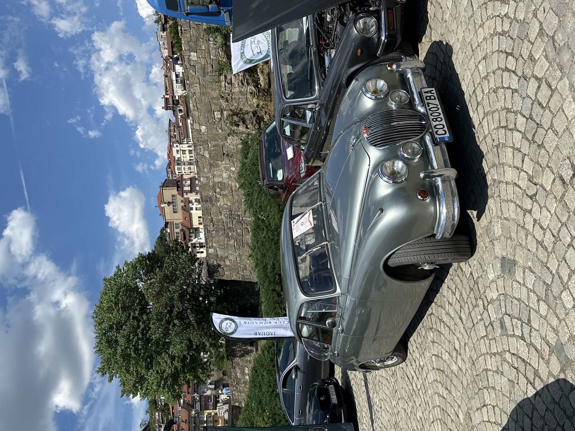 A vintage silver Jaguar Mk2 saloon car is parked on a cobblestone street in Veliko Tarnovo, Bulgaria, under a bright blue sky with scattered clouds. The car, with its distinctive grille and chrome accents, is the primary focus. Behind it, another car, possibly a Jaguar XJ, has its hood open, revealing its engine. Further in the background, a red sedan is visible. Two large flags, one white with a green Jaguar logo and the text "JAGUAR CLUB ROMANIA," are planted behind the cars. The backdrop features a stone wall, lush green bushes, and a panorama of traditional Bulgarian houses with red-tiled roofs climbing a hillside, characteristic of Veliko Tarnovo's historic architecture. Two people, one wearing a blue shirt and another in a white cap, are casually observing the scene in the distance. The overall setting suggests an outdoor car show or gathering in a historically rich urban environment.