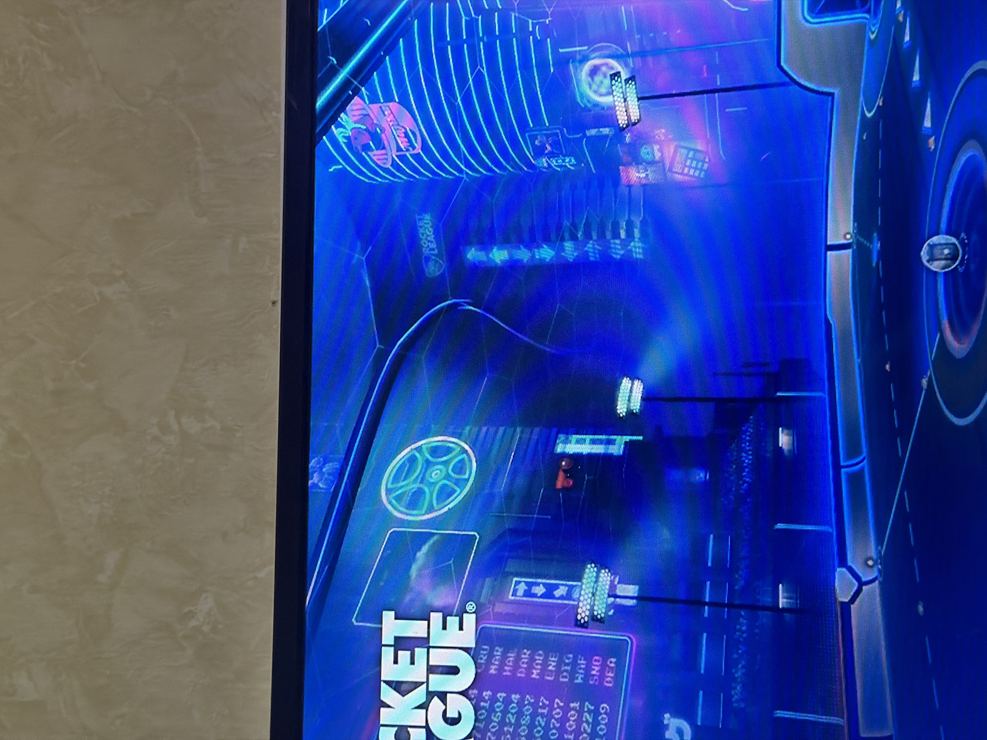 The image captures a vertically oriented display screen, likely a television or monitor, showing a video game. The screen occupies the right half of the frame, bordered on its left by a light-colored, textured wall. The game depicted is "Rocket League," as indicated by the prominent, rotated logo "ROCKET LEAGUE" visible in the bottom-left quadrant of the screen.
The game environment is a futuristic, neon-lit arena rendered predominantly in vibrant blue and purple tones, with glowing linear patterns outlining structures and pathways. Various abstract game elements are scattered throughout the scene, including small representations of cars, glowing boost pads, and hexagonal floor patterns. A circular emblem is visible near the center-left of the screen, and what appears to be a scoreboard or statistics panel is displayed in the bottom-center.
On this panel, a list of alphanumeric data is present, featuring scores or identifiers alongside three-letter abbreviations:
- 71014 CRU
- 70604 HAR
- 51204 HAL
- 60807 DAR
- 60217 MOD
- 0207 ENE
- 1001 DIG
- 0227 HAF
- 1009 SNO
- 1009 DEA
The off-screen area reveals a beige or light brown wall with a subtle marbling or textured pattern, suggesting an indoor setting where the photograph was taken. The photograph itself is rotated 90 degrees clockwise, making the screen appear as if it's placed vertically on its side in the image. No specific time of day or weather details are discernible from the content.
