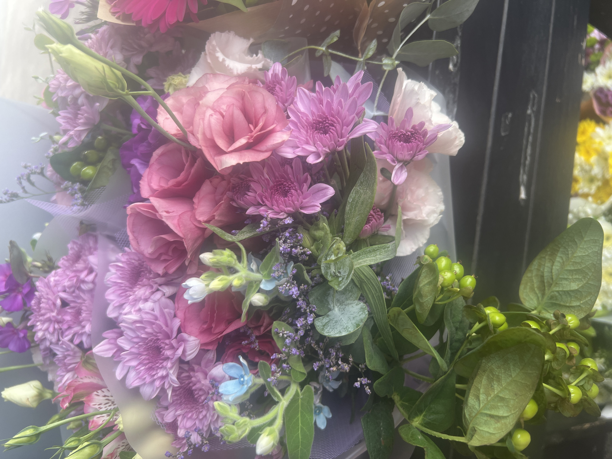 A vertical, close-up, eye-level shot captures a bouquet of vibrant flowers. The bouquet is predominantly filled with pink roses and purple chrysanthemums, interspersed with delicate blue and white blossoms and sprigs of tiny purple flowers. Green foliage and branches with small, green berries add a natural touch to the arrangement. The flowers are partially wrapped in translucent purple and white paper, adding a soft, diffused quality to the image. The background is blurred, with hints of other flowers and a dark, reflective surface. The overall lighting is soft and diffused, creating a dreamy and ethereal effect. The image is taken in Hadera, Israel.