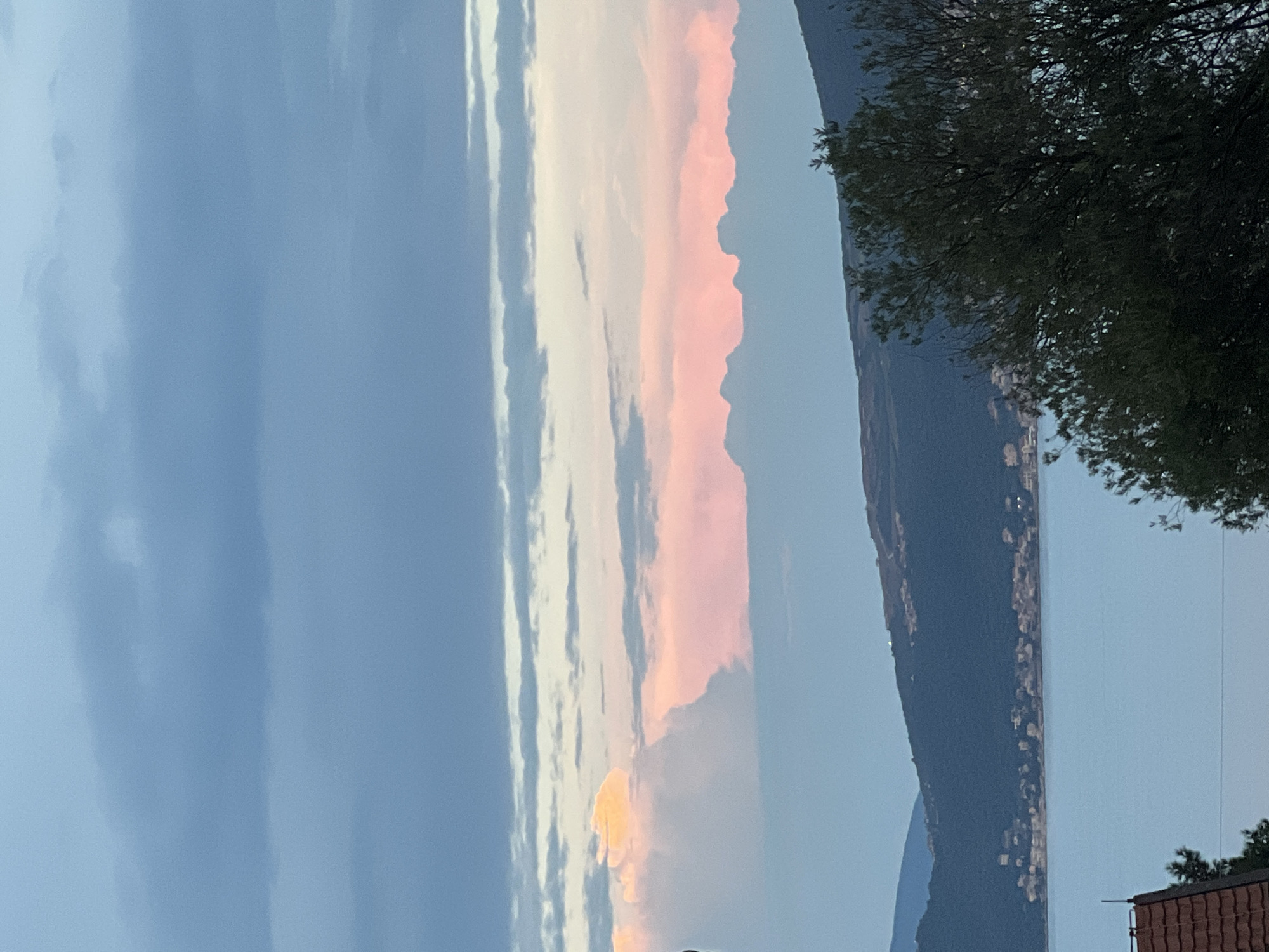The image captures a panoramic view of Baošići, Montenegro, during what appears to be twilight. The sky is a dominant feature, displaying layers of clouds in various shades of blue and gray, with streaks of soft pink and yellow indicating the lingering light of sunset or sunrise. Below the sky, a range of dark, forested hills slopes down towards a body of calm, light blue water. Along the shoreline of the hills, a scattering of lights and buildings suggests a settlement within the town. In the foreground, the dark silhouette of trees frames the scene, with a portion of a reddish-brown tiled roof visible in the lower left corner, hinting at residential architecture. The overall impression is one of serene natural beauty, with a human presence subtly integrated into the landscape.