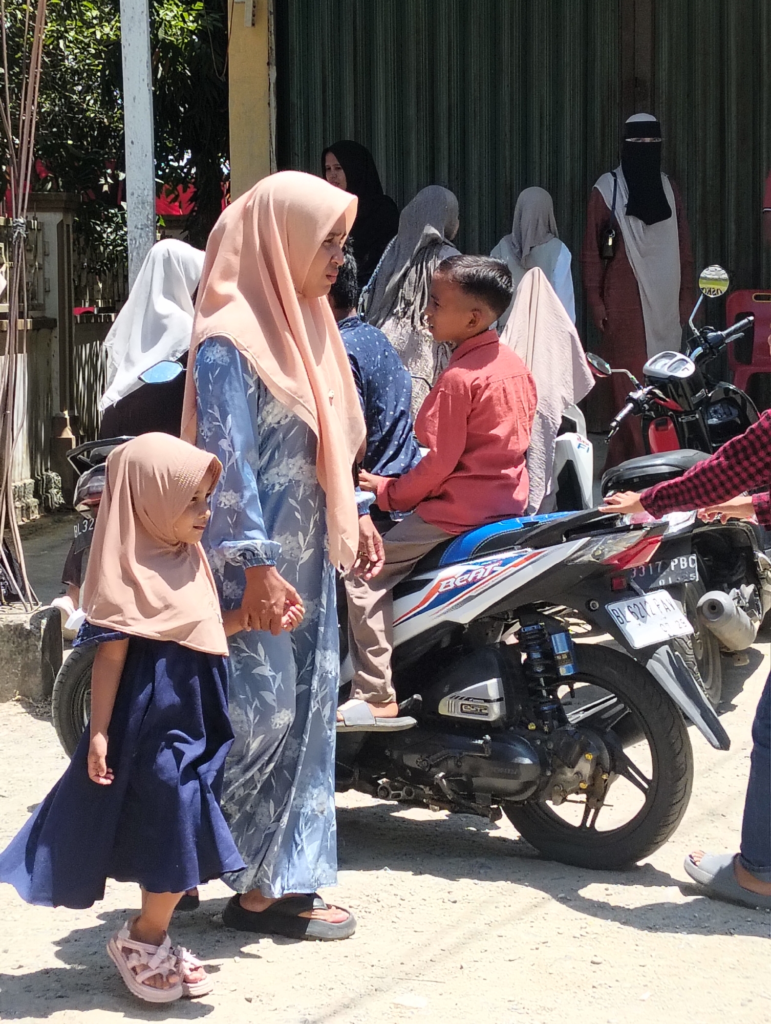 In the city of Teungoh, Indonesia, under a clear sky, a group of people are gathered outdoors, suggesting a public space like a street or a marketplace. The focal point is a young girl in a light brown hijab and a dark blue dress, holding hands with an adult woman wearing a light pink hijab and a blue floral patterned dress. The woman is looking to her right. Behind them, other individuals are visible, some in traditional Islamic attire. A boy in a coral-colored shirt sits on a white and blue motorcycle. Another motorcycle, dark red and black, is parked to the right, with a person wearing a red and black checkered shirt leaning towards it. The ground is a dirt or gravel surface, and a building with green corrugated metal siding is visible in the background. The scene appears to be a casual social gathering, possibly on a warm day given the attire.