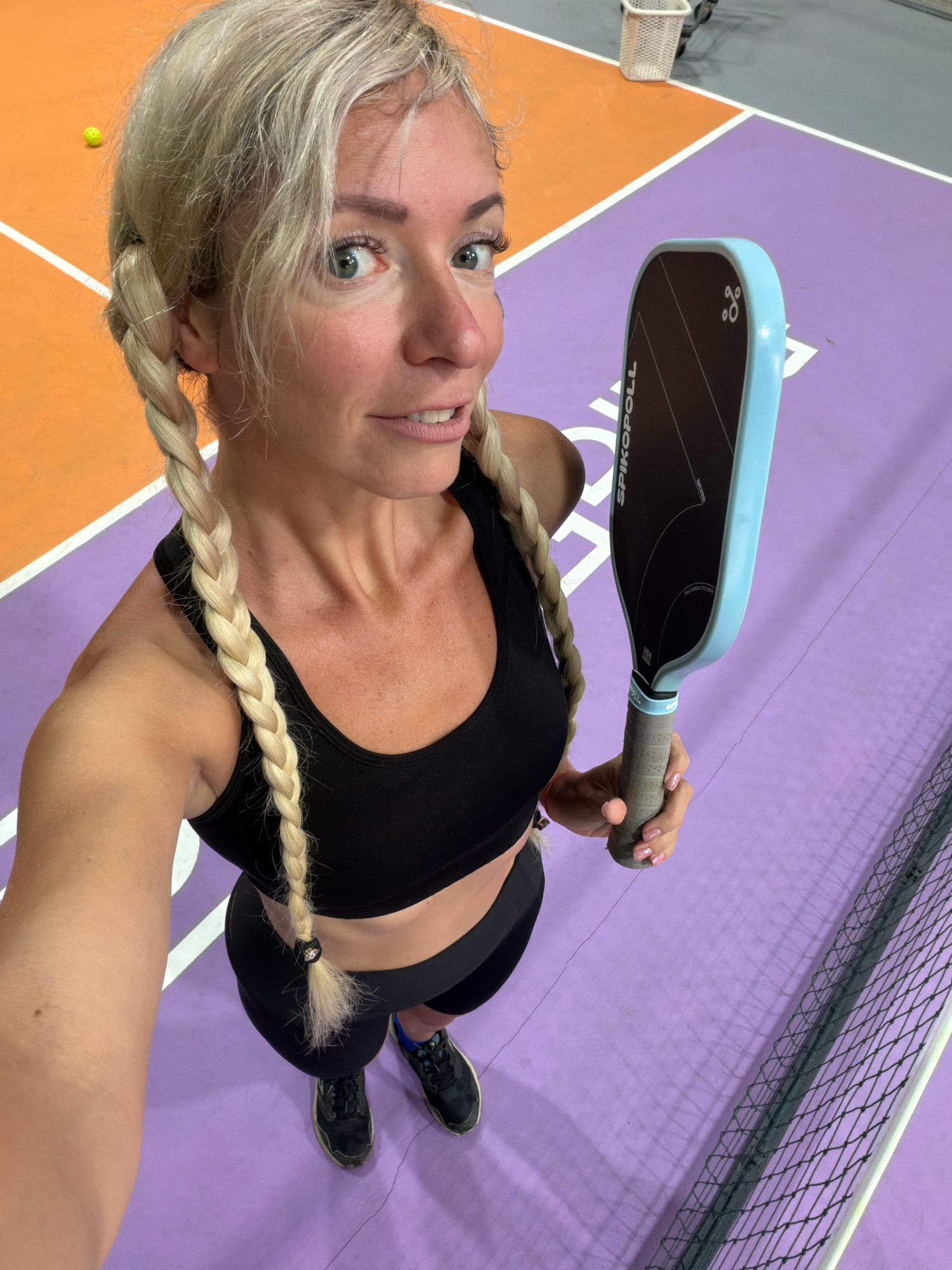 In this high-angle, selfie-style photograph taken on a pickleball court, a blonde woman with braided pigtails holds a light blue pickleball paddle. She is wearing a black sports bra and black athletic shorts, with black tennis shoes on her feet. The court surface is a vibrant purple with white lines marking the boundaries, and an orange section is visible in the background. A yellow pickleball can be seen on the orange section of the court. A white mesh net is present in the lower right corner of the image. The overall scene depicts a sporty environment, likely an indoor or outdoor recreational facility. The image does not provide any information to suggest it was taken in Da Lat, Vietnam.