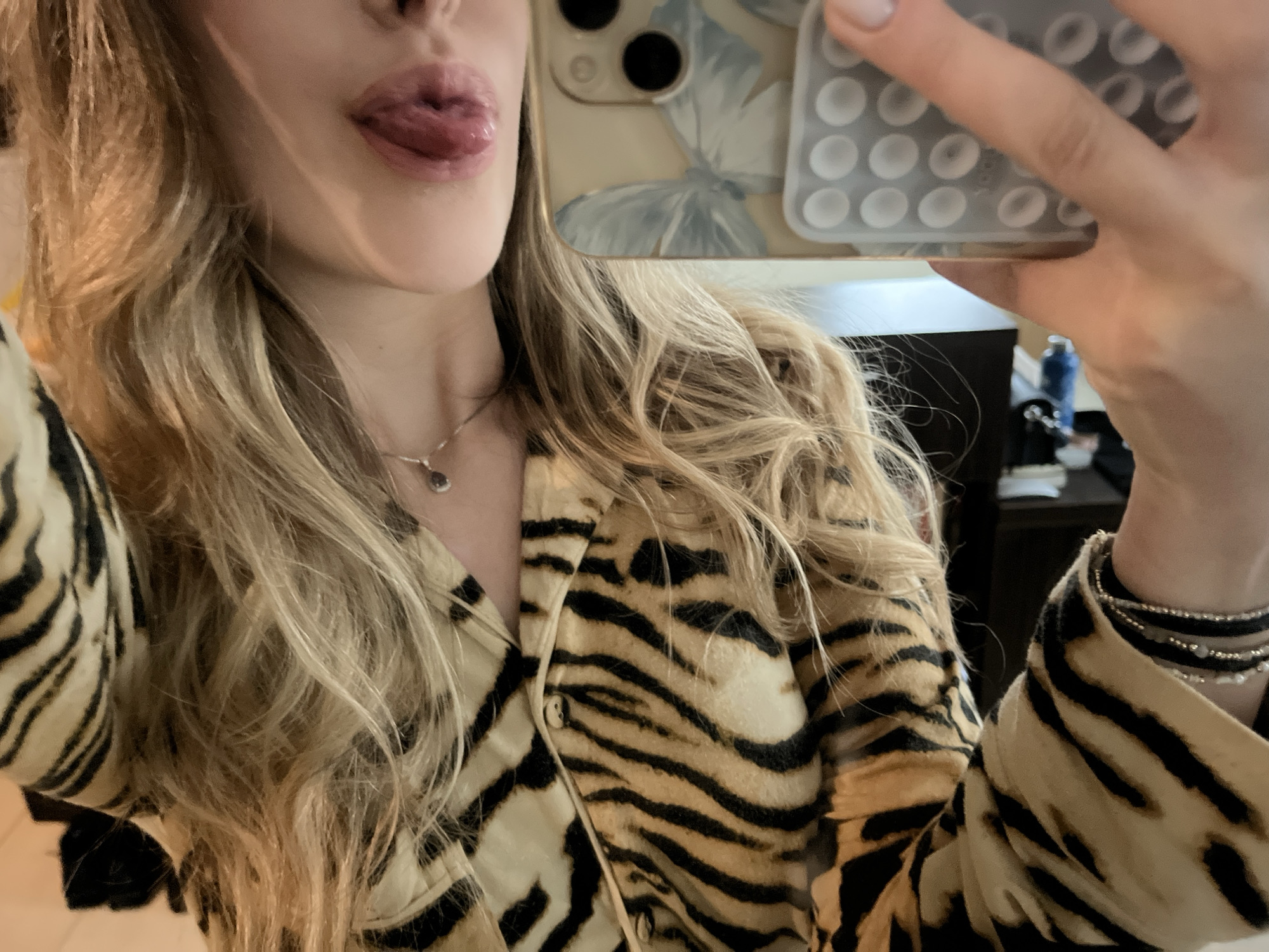 A young, light-skinned woman is taking a selfie. She is wearing a tiger-striped blouse, a delicate necklace, and bracelets on her left wrist. She is sticking her tongue out. She is holding an iPhone in her right hand, with a light-colored case adorned with a butterfly and a "pop it" type accessory. The background is blurry, but appears to be an interior space, possibly a bedroom or living area with dark furniture and bottles on a shelf. There is nothing in the image to indicate the location is Sofia, Bulgaria.