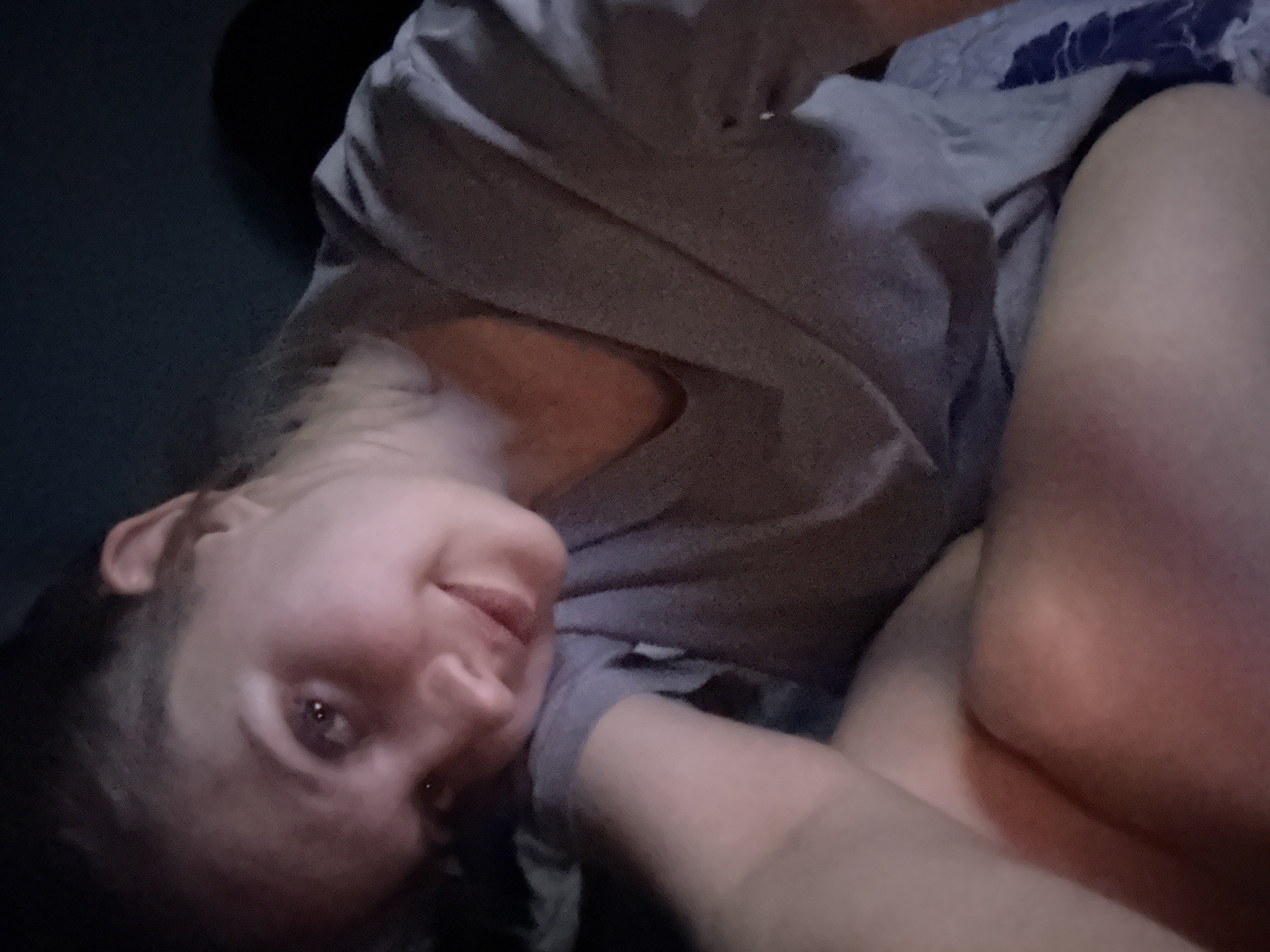 A woman is lying on a surface, possibly a bed or couch, wearing a gray shirt. She is looking at the camera with a slight smile. The background is dark and indistinct. There is no information to indicate this image was taken in Izhevsk, Russia.