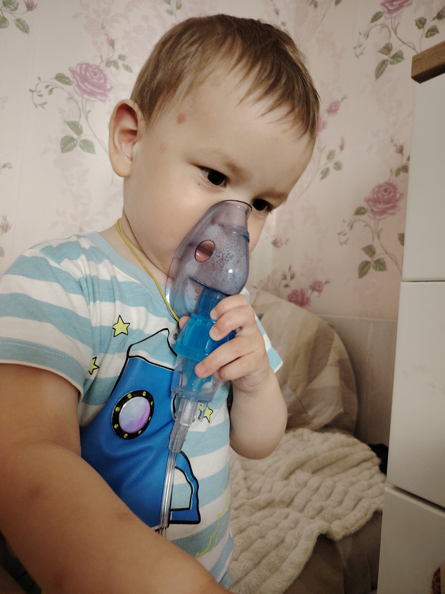 A young child stands holding a blue and clear plastic nebulizer mask to their face. The child wears a light blue and white striped shirt with a blue spaceship design featuring yellow stars. A thin gold chain necklace is visible around the child's neck. The background features light pink floral wallpaper and part of a white cabinet. Based on metadata, this image was taken in Марковский, Russia.
