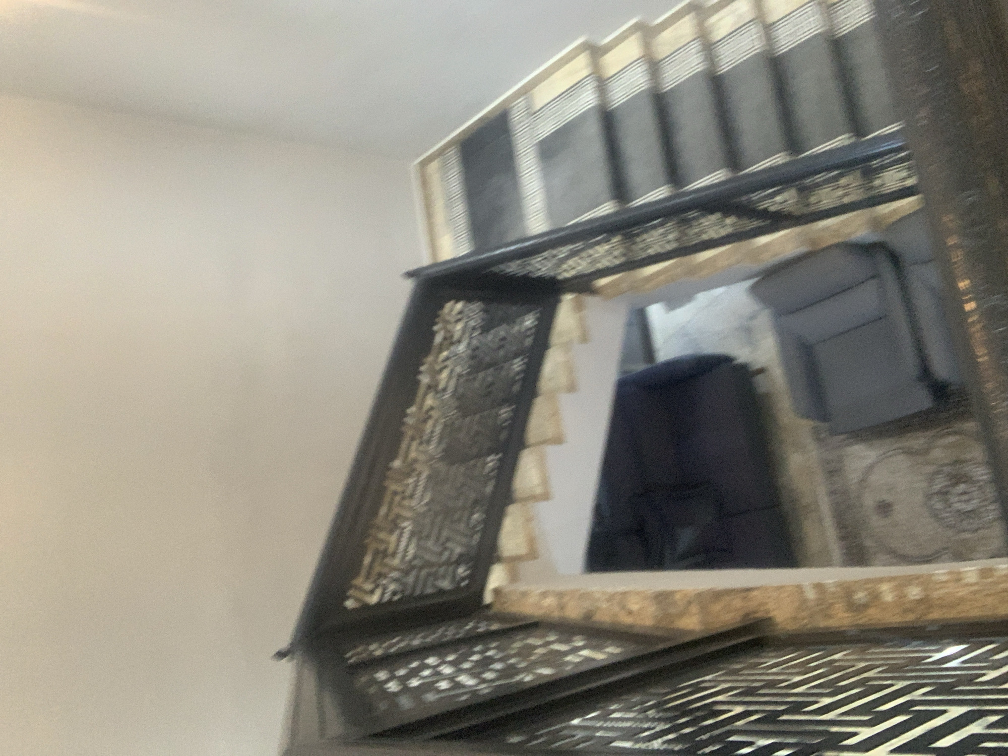 The image shows an interior staircase in Abuja, Nigeria, with a dark, ornate railing and carpeted steps. Below the staircase, a living area is visible, featuring a dark blue sofa and a grey sofa. The walls appear to be a neutral color, and there is a patterned rug on the floor. The lighting suggests it is daytime, with natural light filtering into the space.