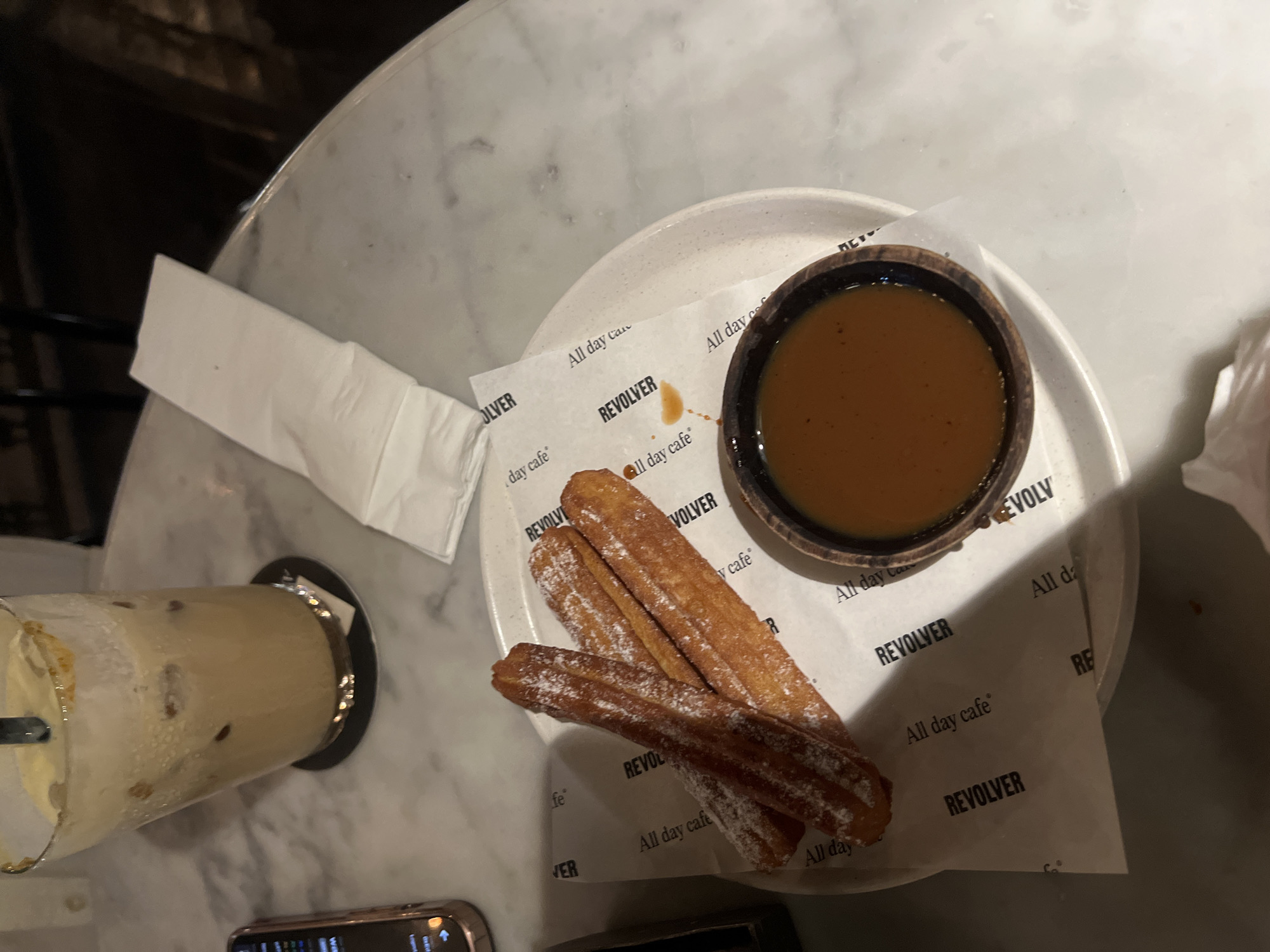 A top-down view shows a dessert of churros with a caramel dipping sauce, accompanied by a tall glass of iced coffee. The churros are dusted with powdered sugar and arranged on a white paper liner featuring the text "REVOLVER All day cafe". The caramel sauce is served in a small wooden bowl. The iced coffee, with ice cubes visible, is in a clear glass with a straw. Both the dessert and the drink are placed on a marble-patterned table. A white napkin is partially visible on the left side of the frame. The lighting suggests it is evening or indoors with low lighting. The scene appears to be in a cafe. The visible text "REVOLVER All day cafe" indicates the name of the establishment.