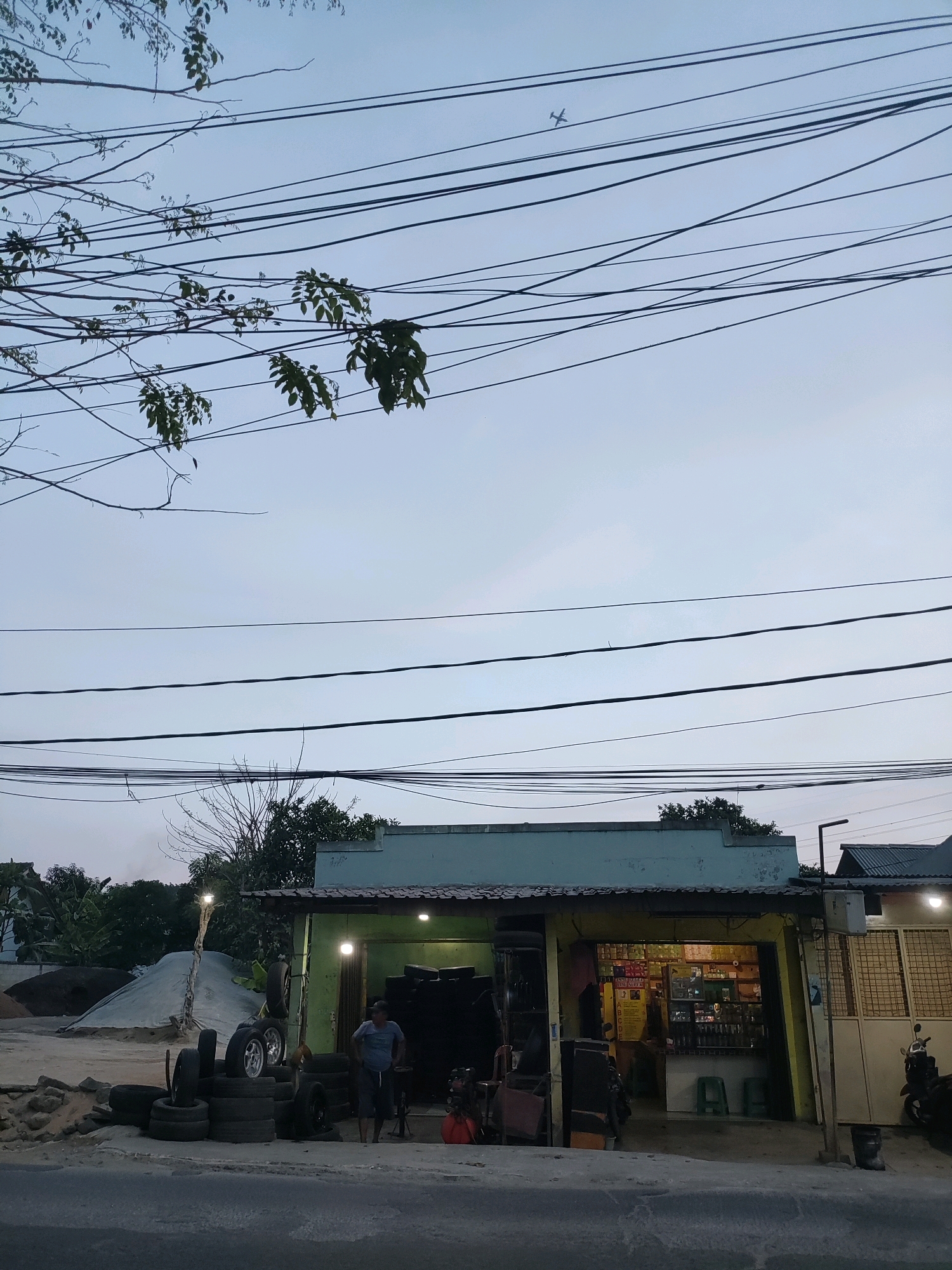 This is a low-angle, eye-level outdoor photograph taken in Wanasari, Indonesia, depicting a roadside tire shop. The sky is a pale blue with a thin layer of clouds, and an airplane is visible in the upper right quadrant. Numerous electrical wires crisscross the frame, some obscured by tree branches in the upper left.
In the foreground, a paved road is visible, with the edge of a sidewalk in front of the shop. To the left of the shop, there are stacks of tires, some arranged in a pyramid shape. Further left, large piles of sand are covered with tarps. A single person, a man wearing a blue t-shirt and dark shorts, stands near the stacks of tires, appearing to be working or overseeing the area.
The shop itself is a simple, open-fronted structure with a green facade and a yellow awning. Inside, shelves are stocked with various items, suggesting it is a small convenience store or a spare parts shop. Two green stools are placed in front of the counter. A motorcycle is parked to the right of the shop. The overall scene suggests a humble, working-class area of Wanasari, likely during the late afternoon or early evening, given the soft light.