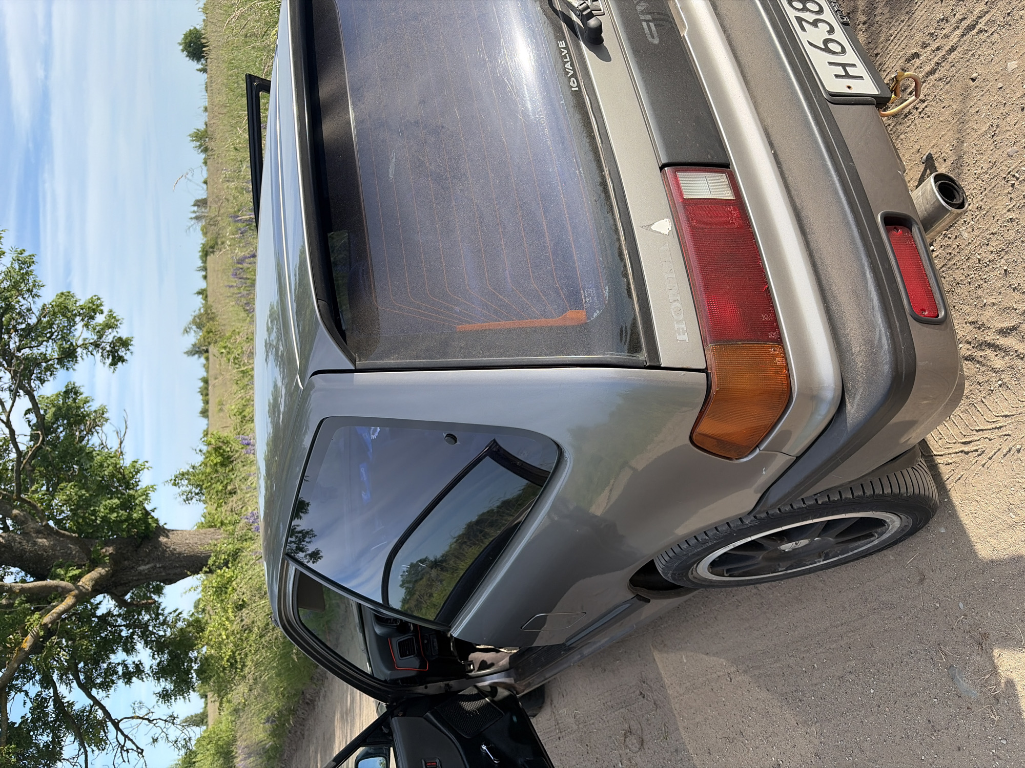 A gray, two-door Honda Civic hatchback is parked on a dirt road, facing right, with its driver's side door open. The car is positioned in a rural landscape with a large tree to the left, green vegetation, and a blue sky with some clouds. The rear of the car is visible, showing a red taillight, a black bumper, a silver exhaust pipe, and a license plate that reads "H638". A small sticker of a white pine tree is visible on the rear of the car. The scene appears to be set in a natural environment, possibly outside the city of Povarovka, Russia, given the context.