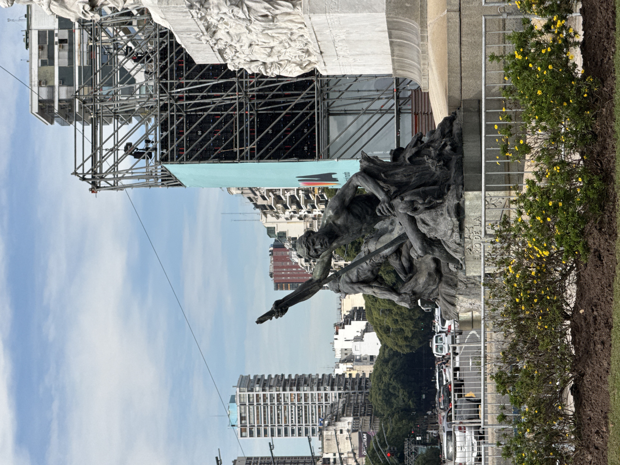 The image depicts a statue with a surrounding cityscape, located in Buenos Aires, Argentina. The statue features human figures in a dynamic pose. Scaffolding and a large screen are positioned behind part of the statue. Buildings of varying heights, a row of trees, and cars on the street are visible in the background. The sky is light blue with clouds. A metal fence and a shrub with yellow flowers are situated near the statue. The image appears to be taken during the day with clear lighting.