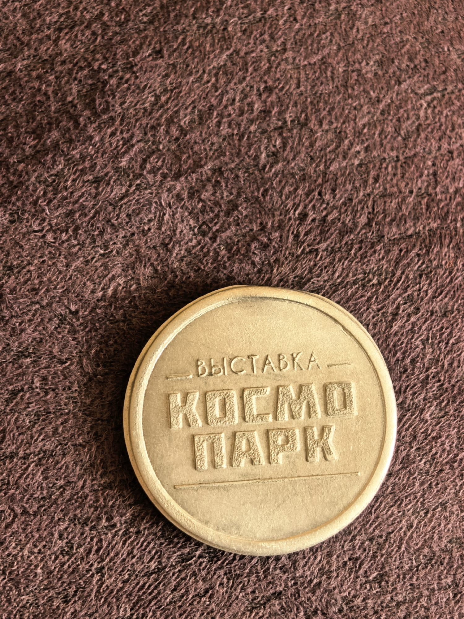 A gold-colored, circular token rests on a textured brown surface. The token has embossed Cyrillic lettering on its face. The top line of text reads "ВЫСТАВКА" (Vystavka), meaning "Exhibition." Below this, the text is split into two lines: "КОСМО" (Kosmo), meaning "Cosmo" or "Space," and "ПАРК" (Park), meaning "Park." The token appears to be an admission ticket or souvenir for an exhibition. The brown surface is likely a fabric, such as a carpet or blanket, with a shaggy texture. There is no indication of any specific location in Sterlitamak, Russia, within the image itself.
