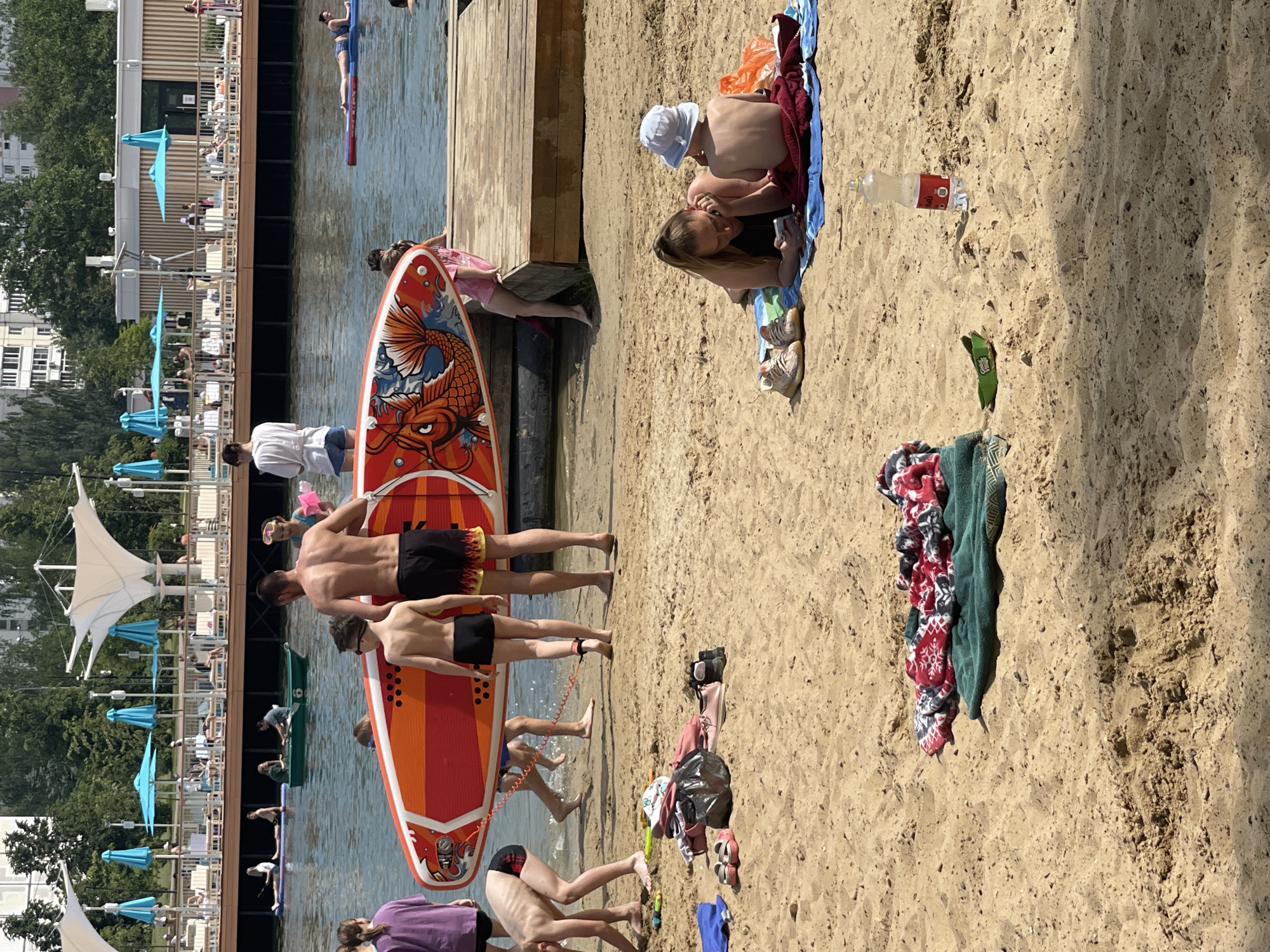 This image captures a sunny, bustling scene at the Northern River Station Park (Severny Rechnoy Vokzal) in Moscow, Russia. The foreground features a sandy beach where multiple individuals are present. On the left, a young person in a purple top and dark shorts stands partially visible. To their right, a young boy with light hair in checkered swim trunks bends over near the water's edge, interacting with the sand. Further right, a man with a muscular back, wearing dark swim shorts with a red and yellow flame pattern, holds a large red and orange paddleboard adorned with a detailed fish design, possibly a koi or dragon. Beside him, a young boy in dark swim trunks stands, an ankle leash from the paddleboard connected to his left ankle. Both the man and boy are oriented towards the calm body of water. A woman in a white top and light shorts observes from slightly behind them, while a young girl in a pink swimsuit with goggles on her head is partially visible to the right of the paddleboard. Scattered on the sand in the mid-foreground are various personal items, including a pile of clothing, a black plastic bag, and sandals. Near the bending boy, yellow and green beach toys are visible. In the middle ground, the calm water shows several activities. A person lies prone on a blue inflatable mat or paddleboard. Further left, another person is on a white paddleboard. A green rowboat, containing two individuals, is also visible. Near the right edge of the frame, a person is partially submerged in the water. Further right on the sand, a woman with blonde hair tied in a high ponytail lies on her stomach on a blue towel, looking at a smartphone. Beside her, a young child, wearing a light blue bucket hat and red swim bottoms, sits facing away on a red towel. Near them, a pair of beige sneakers are placed on the sand. A red and white patterned blanket and a green towel are folded nearby. A clear plastic bottle with a red label stands upright in the sand, next to a green flip-flop. The background reveals a modern recreational complex along the water's edge. An elevated wooden deck, furnished with numerous white lounge chairs and light blue sun umbrellas, extends across the scene. Several white, sail-like tensile fabric canopies provide shade over parts of the deck. People are