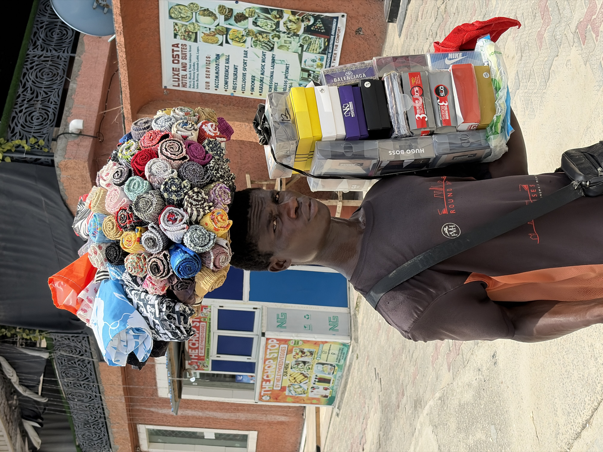 A street vendor is captured in a bright daylight scene in Igboefon, Nigeria, actively engaged in selling goods. The individual, appearing to be male, expertly balances a large, colorful, and intricately arranged mound of rolled fabrics or textiles on his head. In his right hand or supported by his arm, he carries a clear plastic container filled with various packaged items, including what appear to be undergarments and accessories.
The vendor wears a dark t-shirt, which has visible text such as "ROUND" and "FVH," and a black cross-body bag. His expression is neutral as he looks forward. The setting is an urban street under sunny weather, with commercial buildings and storefronts forming the background.
Notable visible text includes a large sign for "LUXE OSTA HOTEL AND SUITES," detailing its services: "ACCOMMODATION," "CONFERENCE HALL," "RESTAURANT," "EXCLUSIVE SPORT BAR," "SNOOKER," "KARAOKE MUSIC NIGHT," "PROFESSIONAL LAUNDRY SERVICE," and "WIRELESS INTERNET." The sign also provides an address in "Jalem Burton, Jahande Lebhi, Lagos," and contact numbers. Another sign for "THE CHOP STOP BY NELEYMIXEDVEGGIES OUR MENU" is visible on a blue-and-white storefront. Brands visible on the merchandise include "HUGO BOSS," "BALENCIAGA," "NIKE," "NOLLYKINGZ," and "PUMA," alongside phrases like "3 BEST FITS BOXERS" and "TWICE THE COMFORT." Other texts such as "NG," "RISON," and "INGWALI MENHI" are also present.