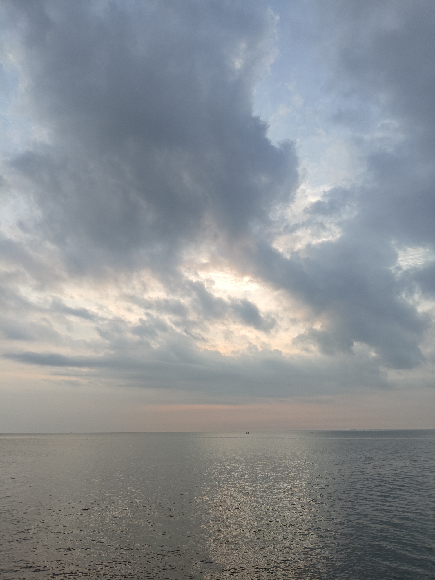 This media file captures a serene seascape under a heavily clouded sky in Lhokseumawe, Indonesia. The image is divided horizontally, with the upper two-thirds dominated by the sky and the lower third by the calm water.
The sky is filled with voluminous, dark grey clouds, ranging from deep charcoal to lighter shades of grey. Patches of lighter, almost pale blue sky are visible between the clouds, particularly towards the upper left. A significant break in the cloud cover in the center allows for a muted, soft light to emanate, suggesting either sunrise or sunset. This light illuminates the edges of the surrounding clouds with subtle hints of warm, pale yellow and orange tones, creating a dramatic and atmospheric effect.
Below the horizon, the sea stretches out, appearing calm with gentle ripples visible on its surface, especially in the foreground. The water reflects the muted light from the sky, creating subtle variations in its greyish-blue hue. The horizon line is flat and clear, separating the vast expanse of water from the sky. On the distant horizon, several small, dark silhouettes of what appear to be fishing boats or other small vessels are sparsely scattered, adding a sense of scale and life to the otherwise expansive and tranquil scene.
The overall impression is one of peaceful solitude and natural grandeur, characterized by the interplay of light and shadow under a dramatic sky. There is no visible text in the image.