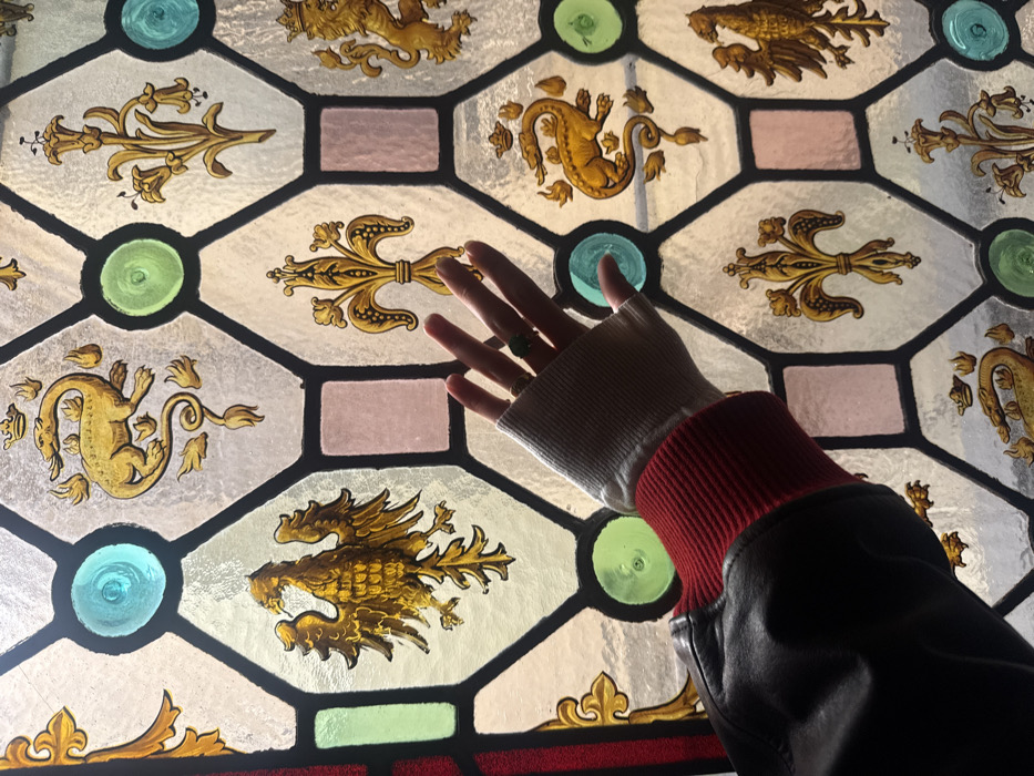 The image displays a detailed section of a stained glass window, identified as being within the Musée Carnavalet, located in the Marais district of Paris, France. The window consists of numerous leaded glass panes forming a geometric pattern of octagonal and rectangular segments. These segments feature various heraldic designs executed in gold and brown paint on clear glass, including multiple depictions of the crowned salamander, a known emblem of King Francis I of France; stylized eagles; fleur-de-lis symbols; and a crowned lion rampant. Interspersed within the leaded grid are small, translucent circular and rectangular panes of colored glass in shades of turquoise, light green, and light pink. A person's left hand, adorned with a light grey ribbed fingerless glove and a ring with a prominent green stone on the ring finger, extends into the foreground. The hand emerges from a dark-colored garment, likely a jacket, distinguished by a visible red ribbed cuff. The hand is positioned centrally, with fingers slightly spread, in front of the illuminated stained glass. The overall scene is brightly lit from behind the window, indicating natural light passing through the glass.