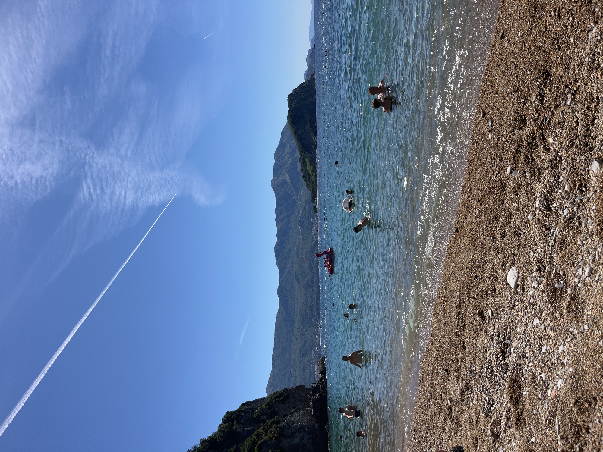 This image depicts a sunny day at a pebble beach in Budva, Montenegro. The foreground is dominated by the textured surface of the beach, with small pebbles and sand. The clear, blue water of the Adriatic Sea extends into the middle ground, where several people are swimming and enjoying the water. Among them, a couple is visible in the right middle ground, facing the viewer. Further out, a pink inflatable flamingo-shaped float with people on it is present in the middle of the sea.
In the background, lush green mountains rise majestically, forming a scenic backdrop to the coastal landscape. A prominent cliff face, covered in vegetation, juts out from the left side of the scene, adding to the dramatic beauty of the bay. The sky is a vibrant blue, with a few wispy clouds and a distinct white contrail from an airplane stretching diagonally across the upper portion of the frame. The overall atmosphere is one of leisure and relaxation on a beautiful summer day.