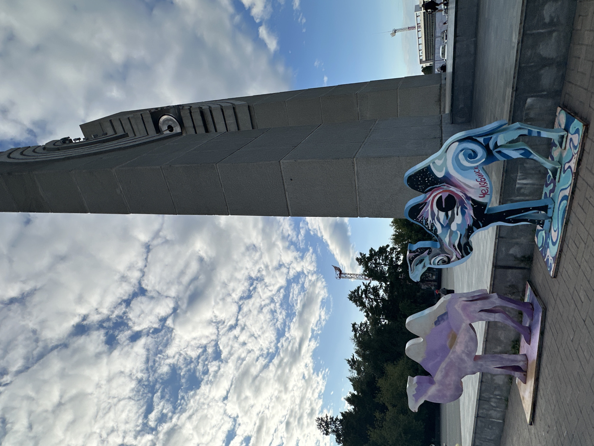 Two stylized, wooden camel sculptures are displayed on a paved plaza. The sculpture on the left is predominantly purple and white, with subtle geometric patterns. The sculpture on the right is adorned with a vibrant, swirling design of blues, blacks, and whites, resembling a starry night sky, with the word "ЧЕЛЯБИНСК" written in cursive on its flank. In the background, a towering, grey, angular monument stands prominently. The sky is filled with scattered white clouds against a bright blue backdrop. A line of trees forms a backdrop to the left, and beyond that, a tall, slender tower with a flag is visible. In the distance, a building with large windows and a light-colored facade can be seen, along with parked vehicles. The plaza pavement is made of grey square tiles.
