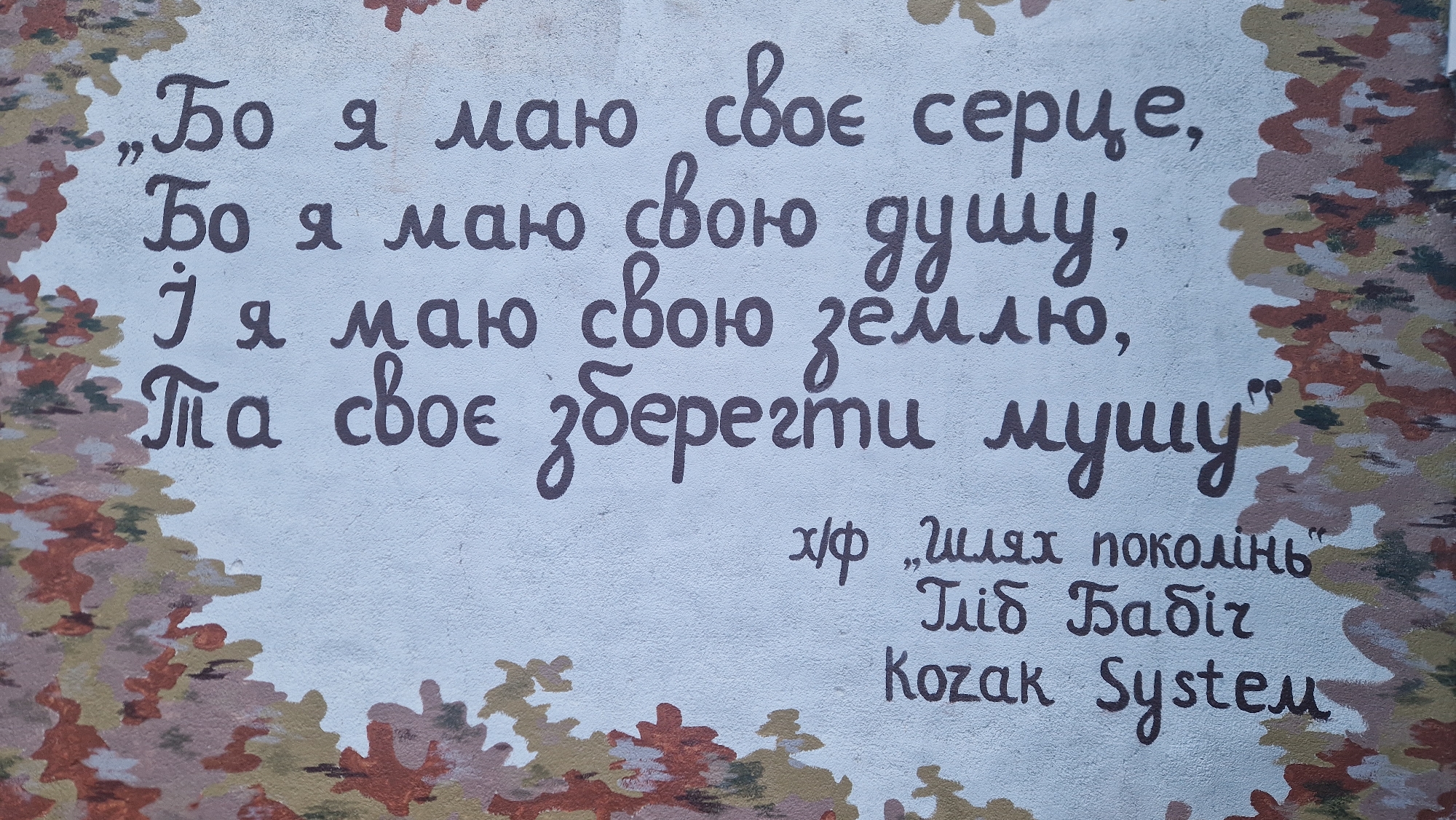 The image shows a section of a light blue wall with a decorative pattern along the bottom and right edges resembling autumn leaves in shades of brown, orange, and green. Handwritten text in dark purple ink is present on the wall. The text is in Ukrainian and appears to be a poem or quote.
The main body of text consists of four lines:
"Бо я маю своє серце,"
"Бо я маю свою душу,"
"Ј я маю свою землю,"
"Па своє зберегти мущу"
Below this main text, there is smaller text on the right side:
"х/ф „шлях поколінь“
ТіГ Бабіг
Kozak System"
The writing style is a decorative script, with some letters elongated and stylized. The overall impression is that of a mural or graffiti art piece. The location is described as Unknown, Unknown, implying no specific geographical information is available.