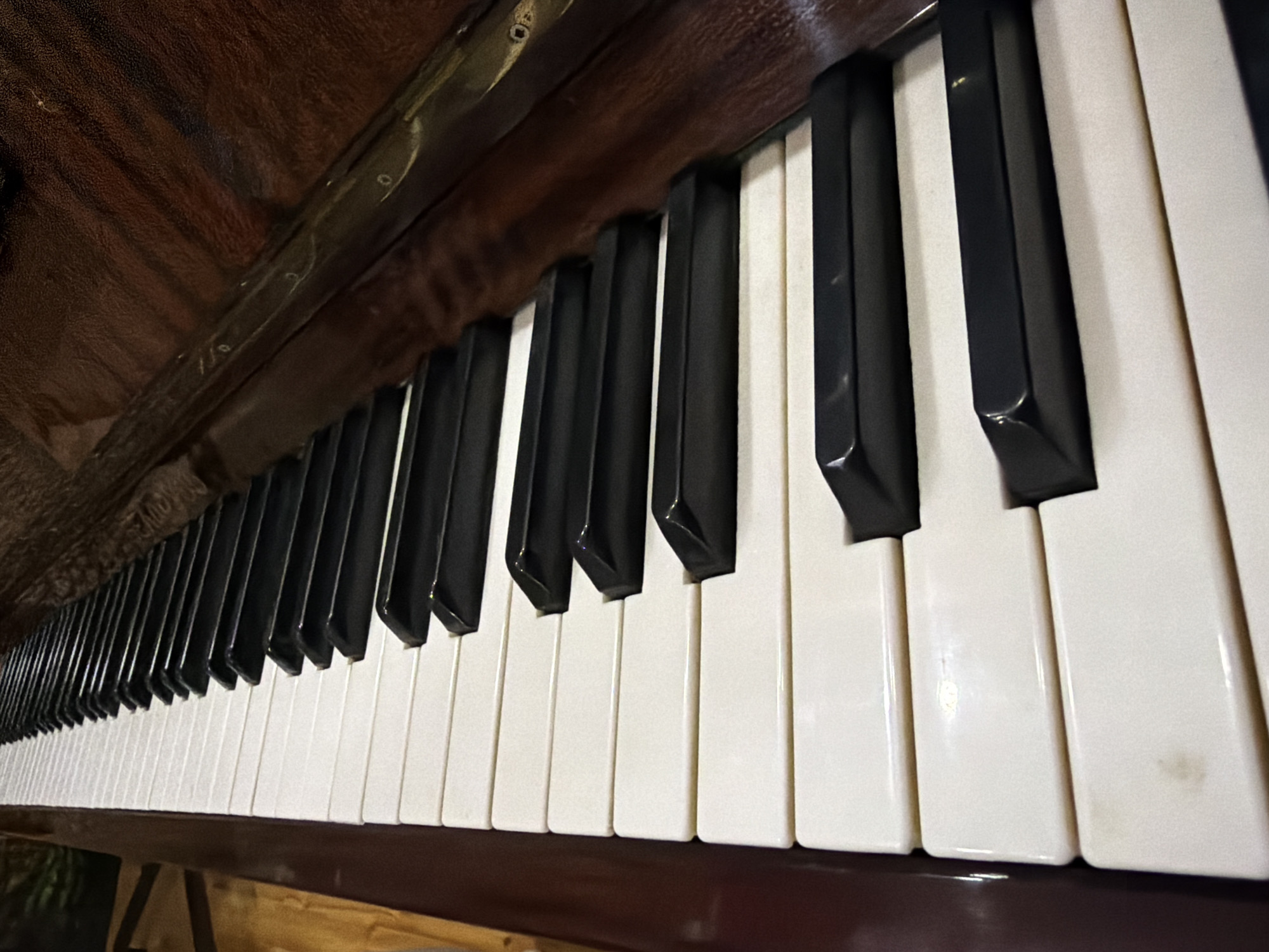 The image displays a close-up, eye-level view of the black and white keys of a piano. The perspective is angled, with the keys extending away from the viewer and towards the upper right of the frame. The piano's wooden casing, a dark reddish-brown, is visible on the right side of the image, with a metallic trim running along the edge of the keys. The lighting suggests an indoor setting, possibly in a home or studio. There are no people or animals present in the image, and no specific action is depicted. The scene is a still life focused on the textures and forms of the musical instrument. The location in the city of Sabirabad, Azerbaijan, cannot be determined from the visual information provided in the image.