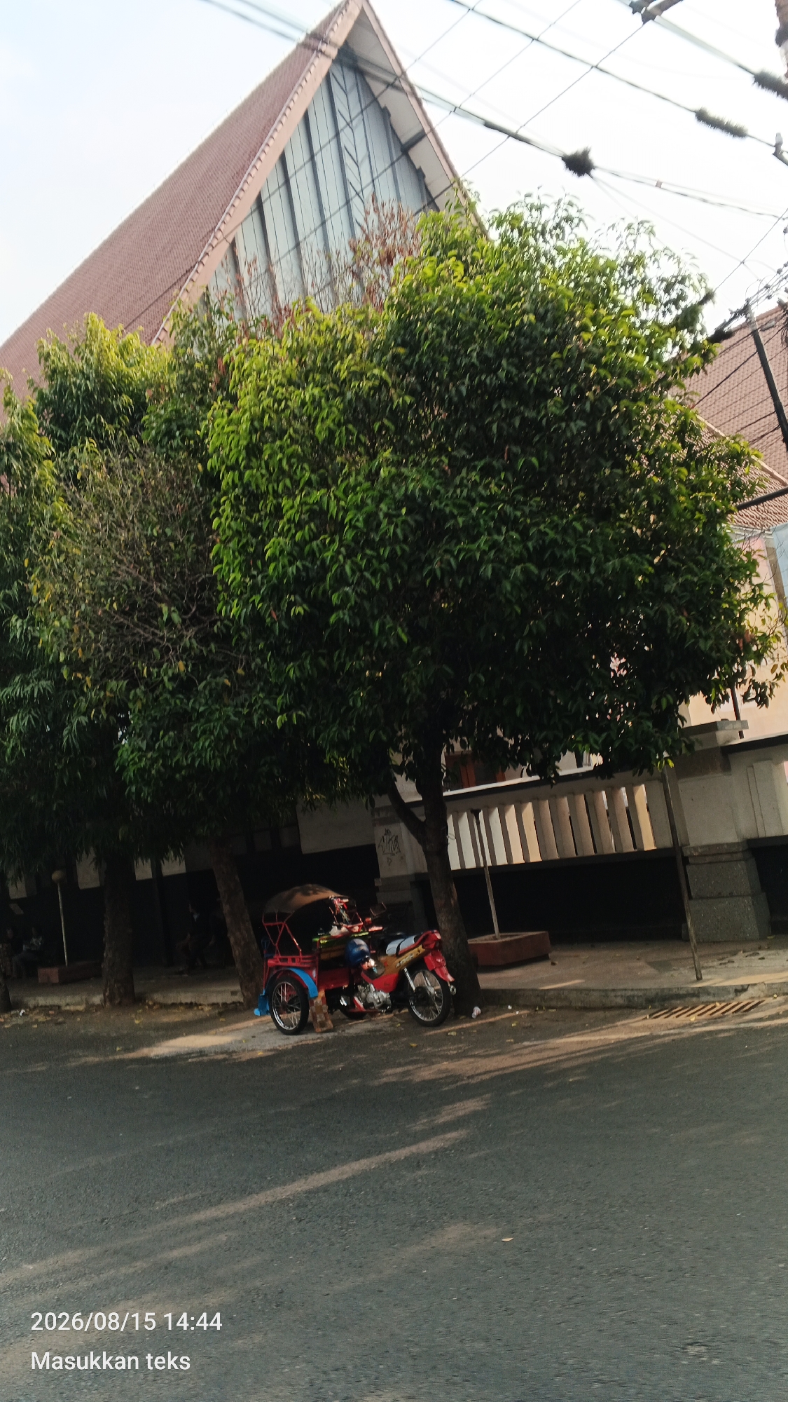 This image captures a street scene in Malang, Indonesia, on August 15, 2026, at 14:44. In the foreground, a paved road is visible, with shadows from trees falling across it. To the right, a red and blue motorized tricycle, commonly known as a bemo or bajaj, is parked by the side of the road, leaning against a tree. The tricycle has a canopy over its passenger area and a single wheel at the front for steering. Behind the tricycle, a building with a distinctive triangular, tiled roof and large glass windows dominates the background. Several lush green trees line the street in front of the building, obscuring some of its lower facade, which appears to have a white railing. The sky is bright, indicating daylight.