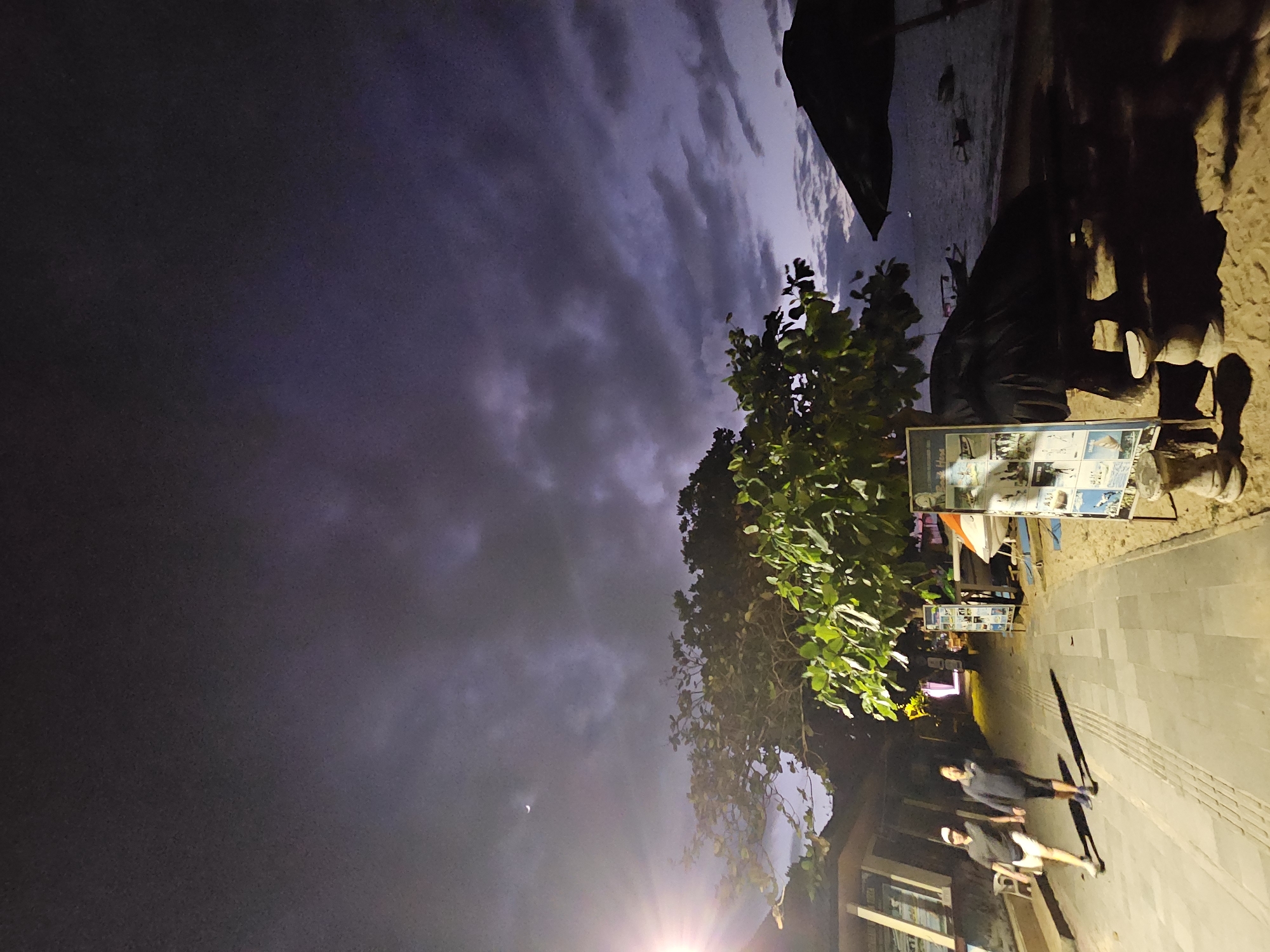A dusk scene on a beach in Denpasar, Indonesia, shows a paved walkway alongside a sandy beach. Two individuals walk on the walkway, one wearing a dark t-shirt and white shorts, the other in a dark jacket and dark shorts. To the left of the walkway, a building with lights is visible, and a bright light source casts a beam to the left. Trees with lush green leaves are situated between the walkway and the beach. On the beach, a large umbrella is open, covering a picnic table. Several informational boards are displayed near the walkway, one of which shows images related to water activities. The sea is visible in the background with a few boats. The sky is dark with dramatic cloud formations and a crescent moon.